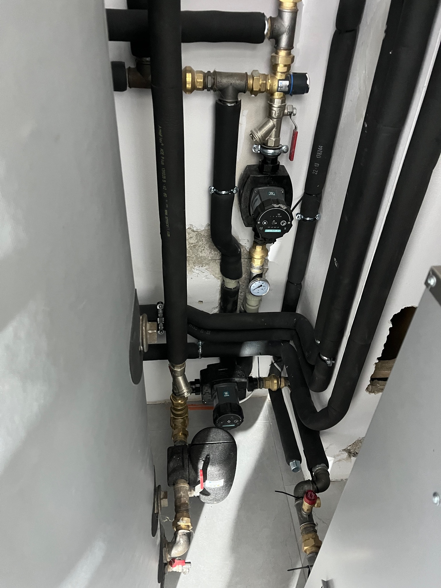 Złożona instalacja grzewcza z czarnymi rurami izolowanymi, zaworami i pompami obiegowymi, zamontowana przy białej ścianie z widocznymi śladami po kuciu.