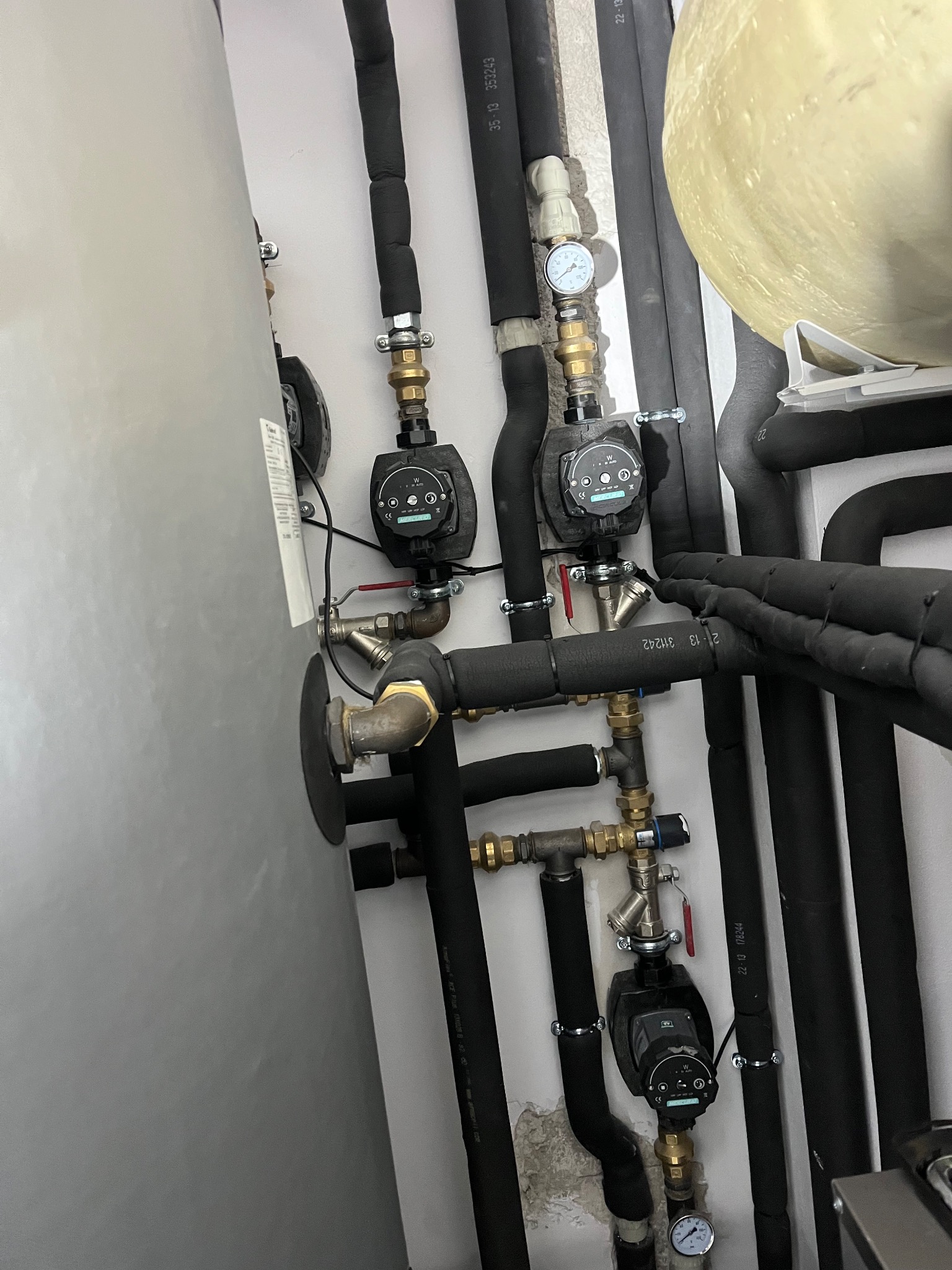 Złożona instalacja grzewcza z rurami w izolacji, pompami obiegowymi i manometrami, zamontowana przy zbiorniku akumulacyjnym w kotłowni. Widoczne liczne złączki i zawory.