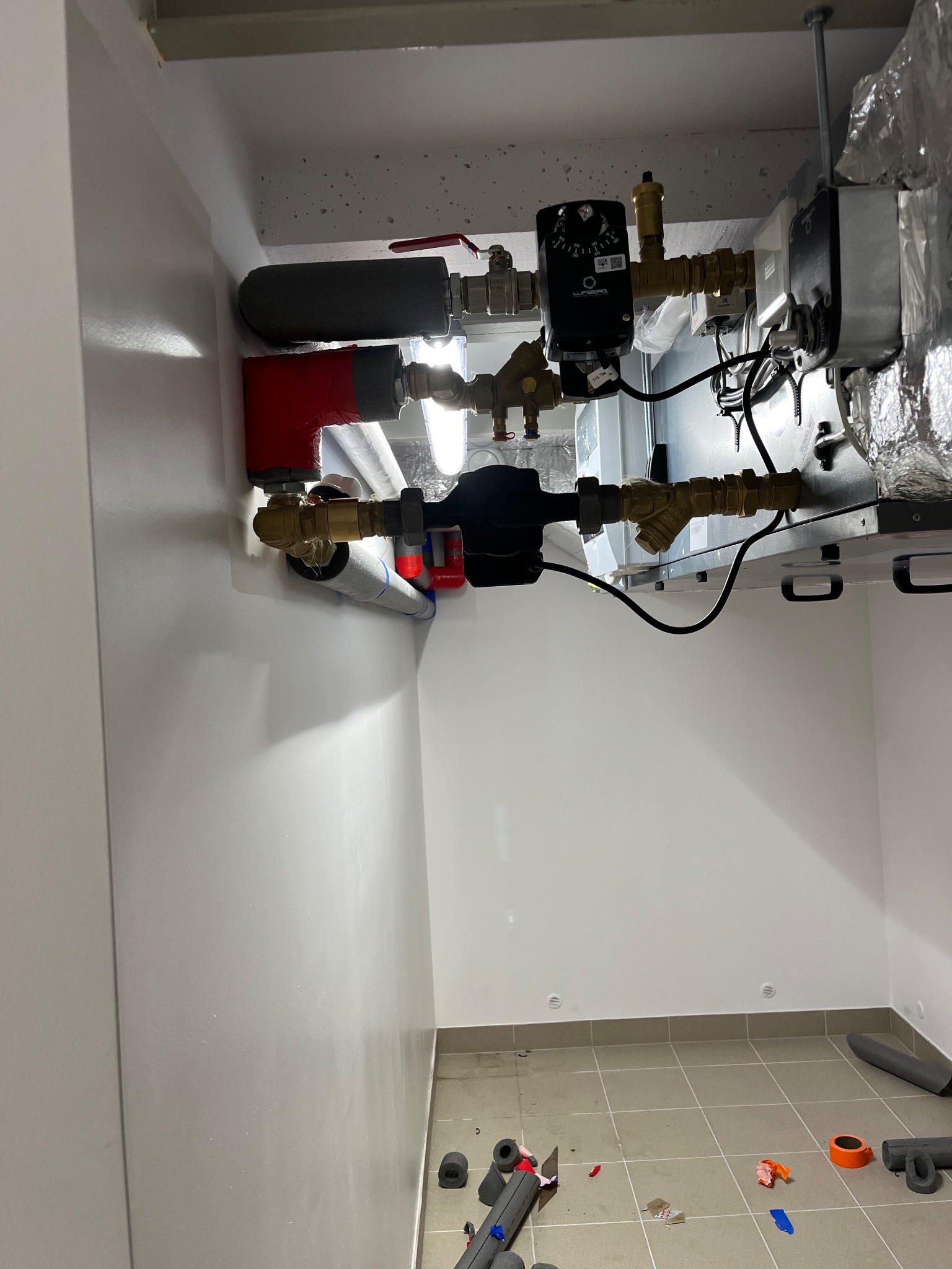 Złożona instalacja grzewcza z widocznymi rurami, zaworami i czarnym regulatorem ciśnienia, zamontowana w białej wnęce. Na podłodze leżą fragmenty izolacji.
