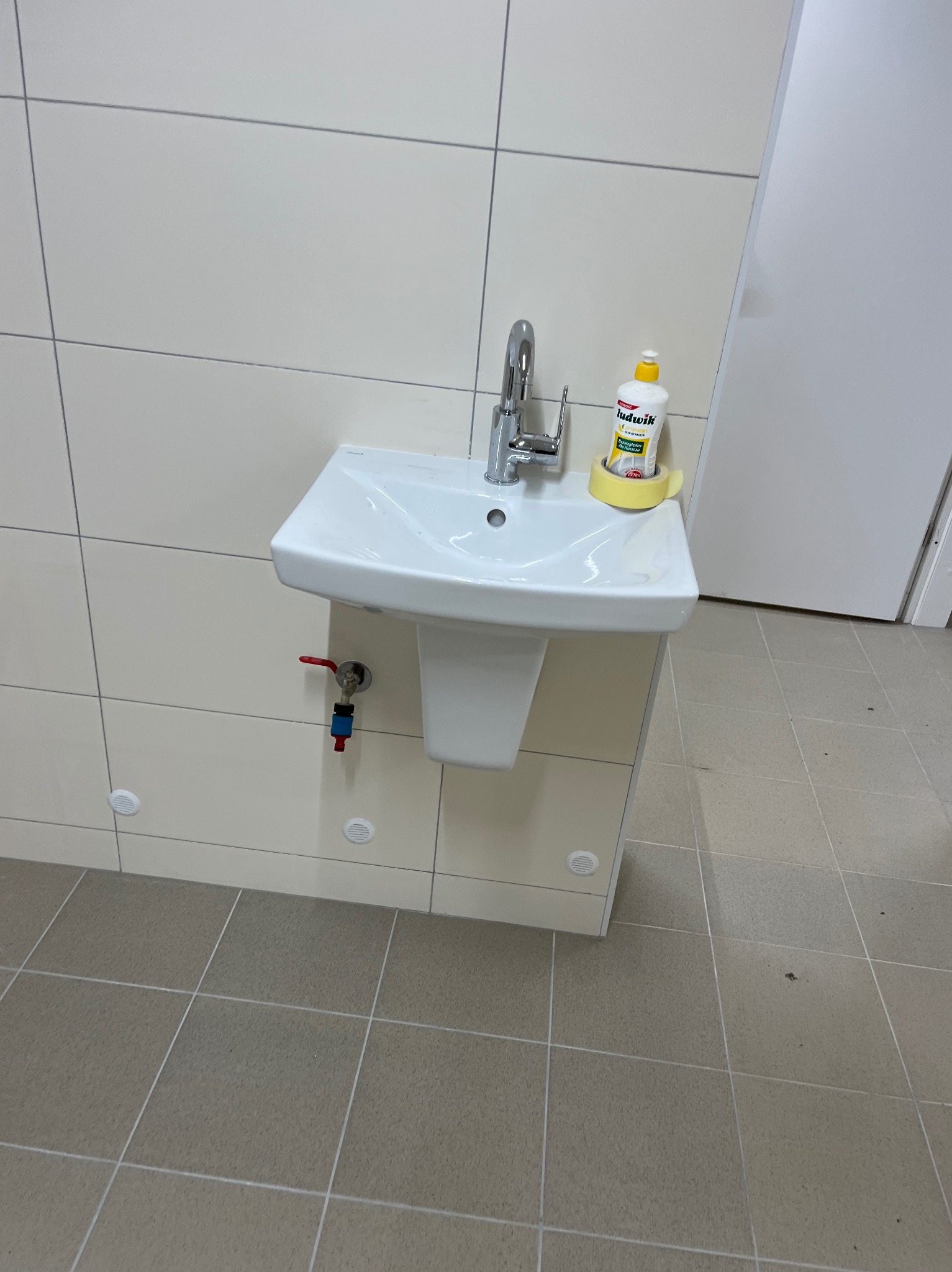 Biała umywalka z chromowaną baterią i zaworem, zamontowana na ścianie z beżowych płytek. Na umywalce płyn do mycia naczyń. Widoczne przyłącza hydrauliczne.