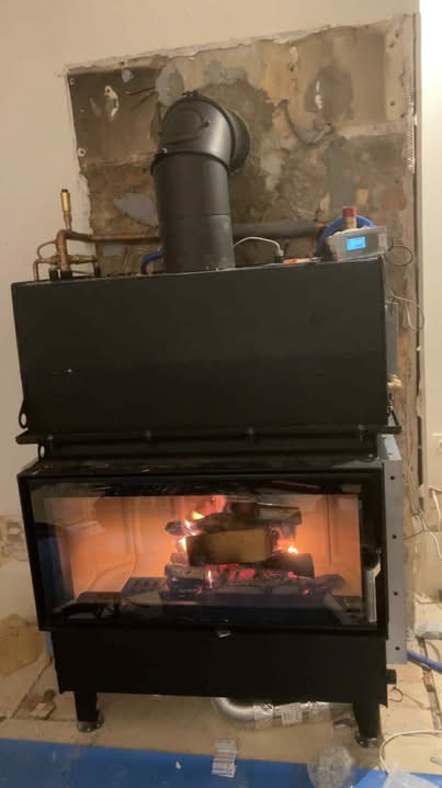 Czarny piec kominkowy z palącym się drewnem, widoczne rury instalacji grzewczej i fragment ściany w trakcie remontu.