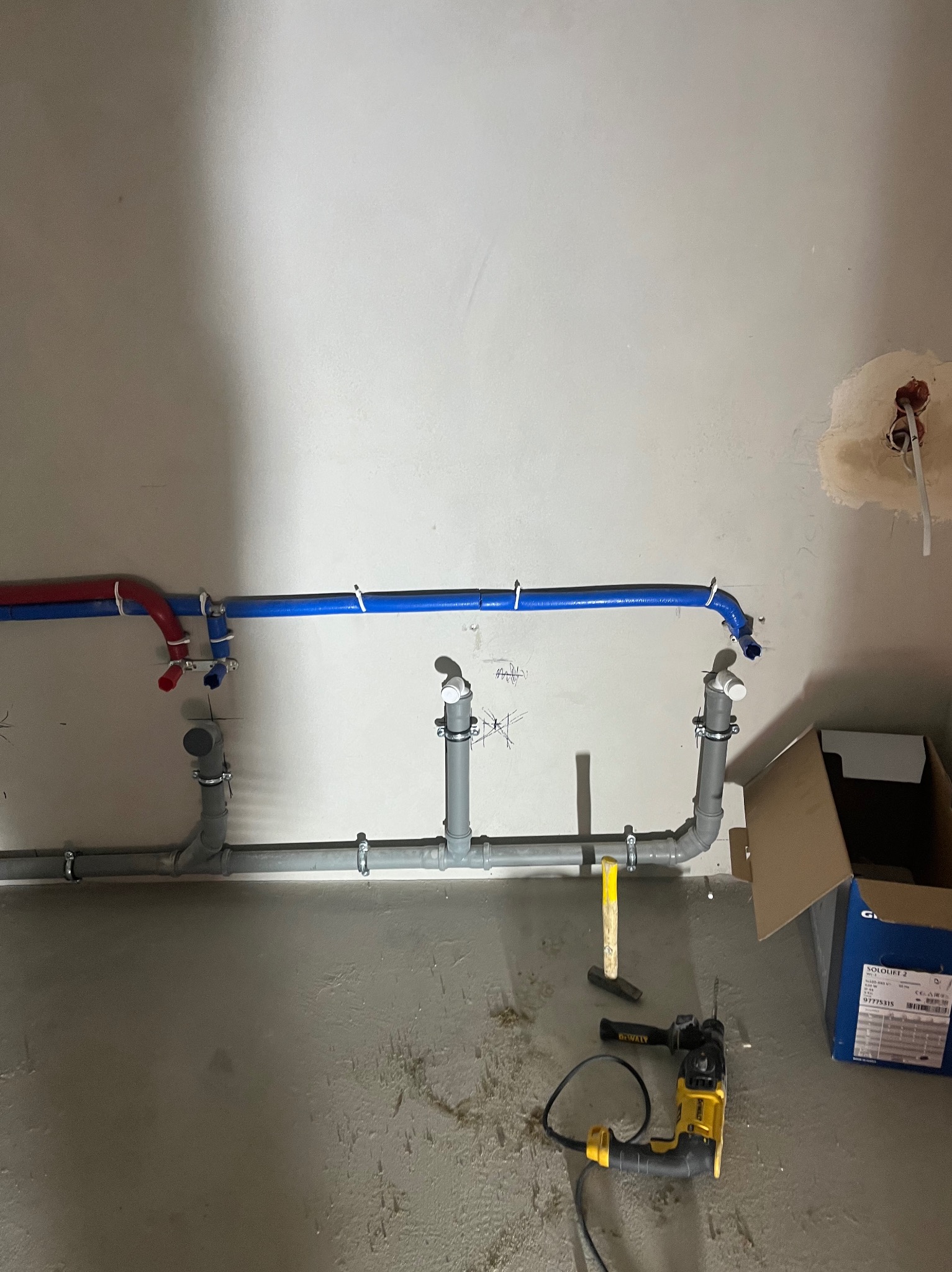 Szare i kolorowe rury instalacji wodnej zamocowane na ścianie, widoczne narzędzia pracy i karton. Instalacja wykonana w surowym pomieszczeniu.
