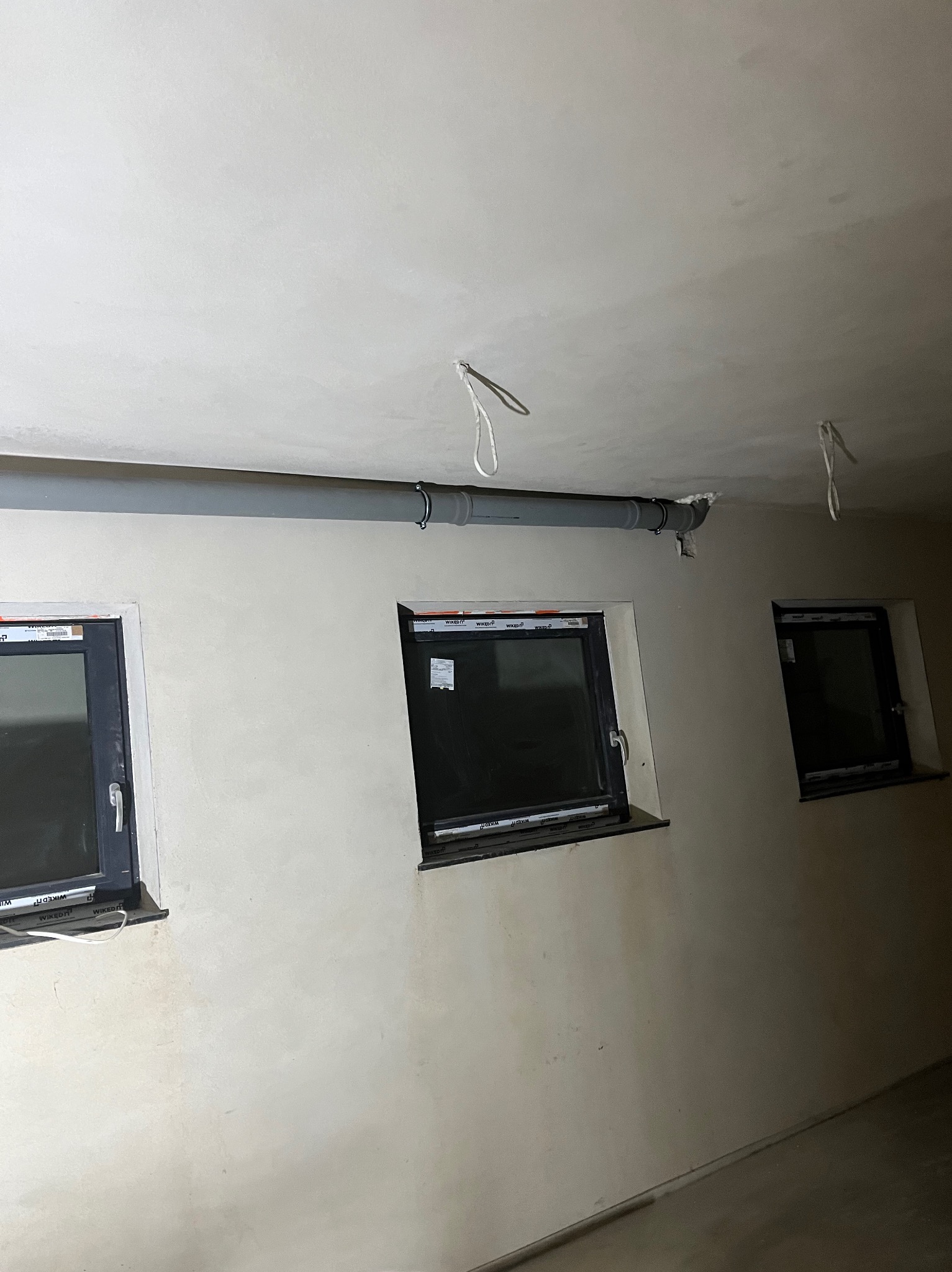 Szara rura instalacyjna zamontowana pod sufitem w pomieszczeniu z trzema oknami o czarnych ramach. Ściany w odcieniach beżu, sufit biały. Widoczne elementy mocujące rurę.