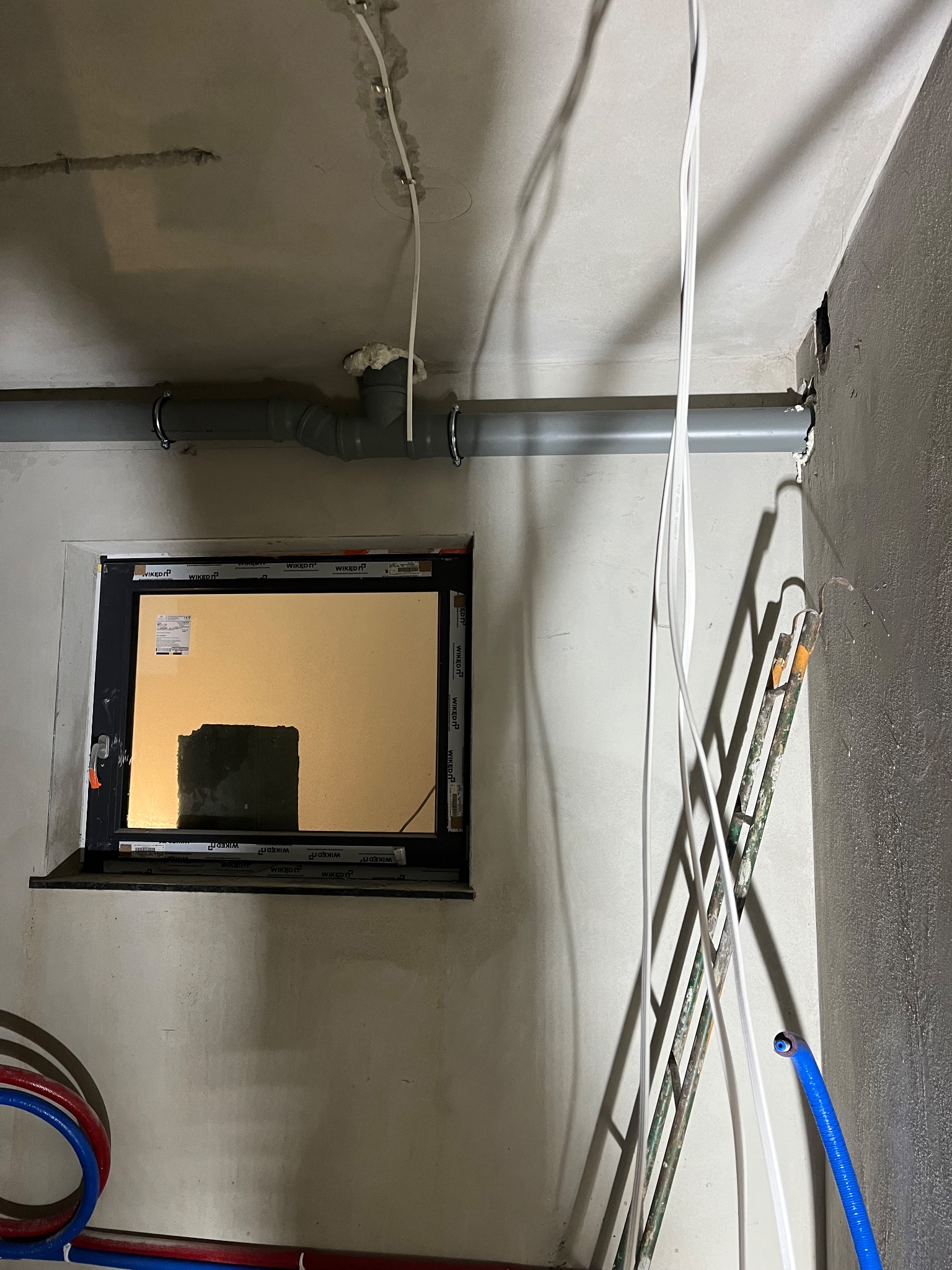Szara rura kanalizacyjna pod sufitem, okno z naklejkami WIKED, kable i drabina w surowym wnętrzu. Instalacja w trakcie prac wykończeniowych.