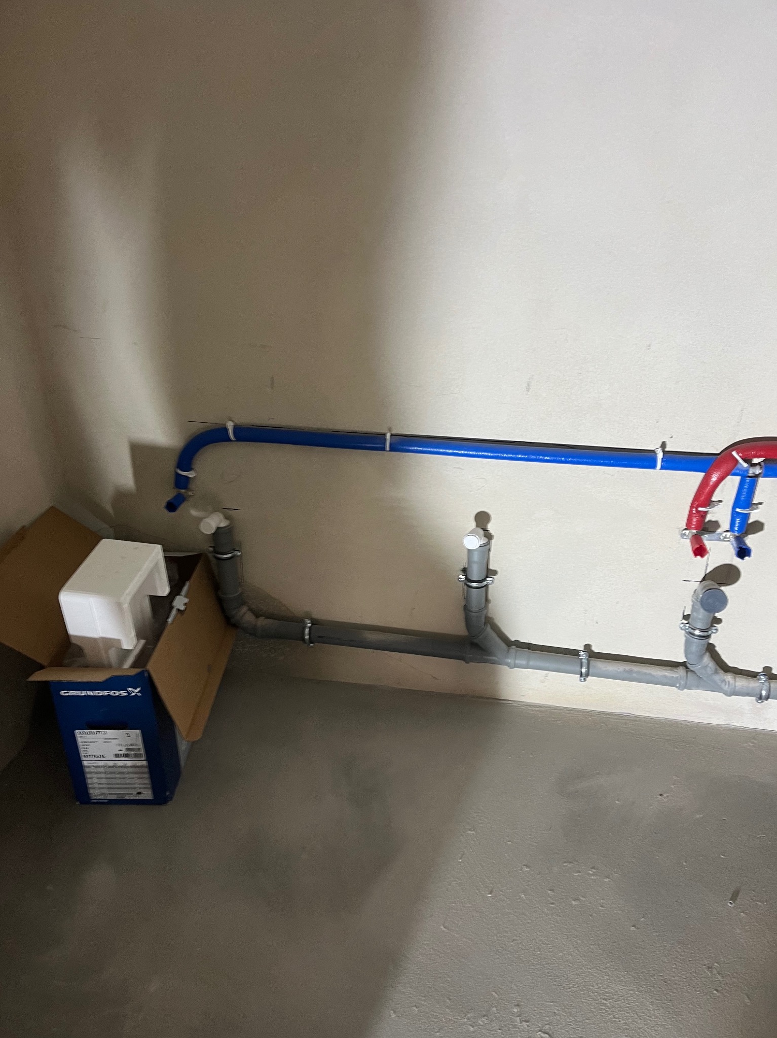 Szare i niebieskie rury instalacji hydraulicznej zamocowane do ściany, obok karton z pompą Grundfos. Widok na fragment instalacji w trakcie montażu.