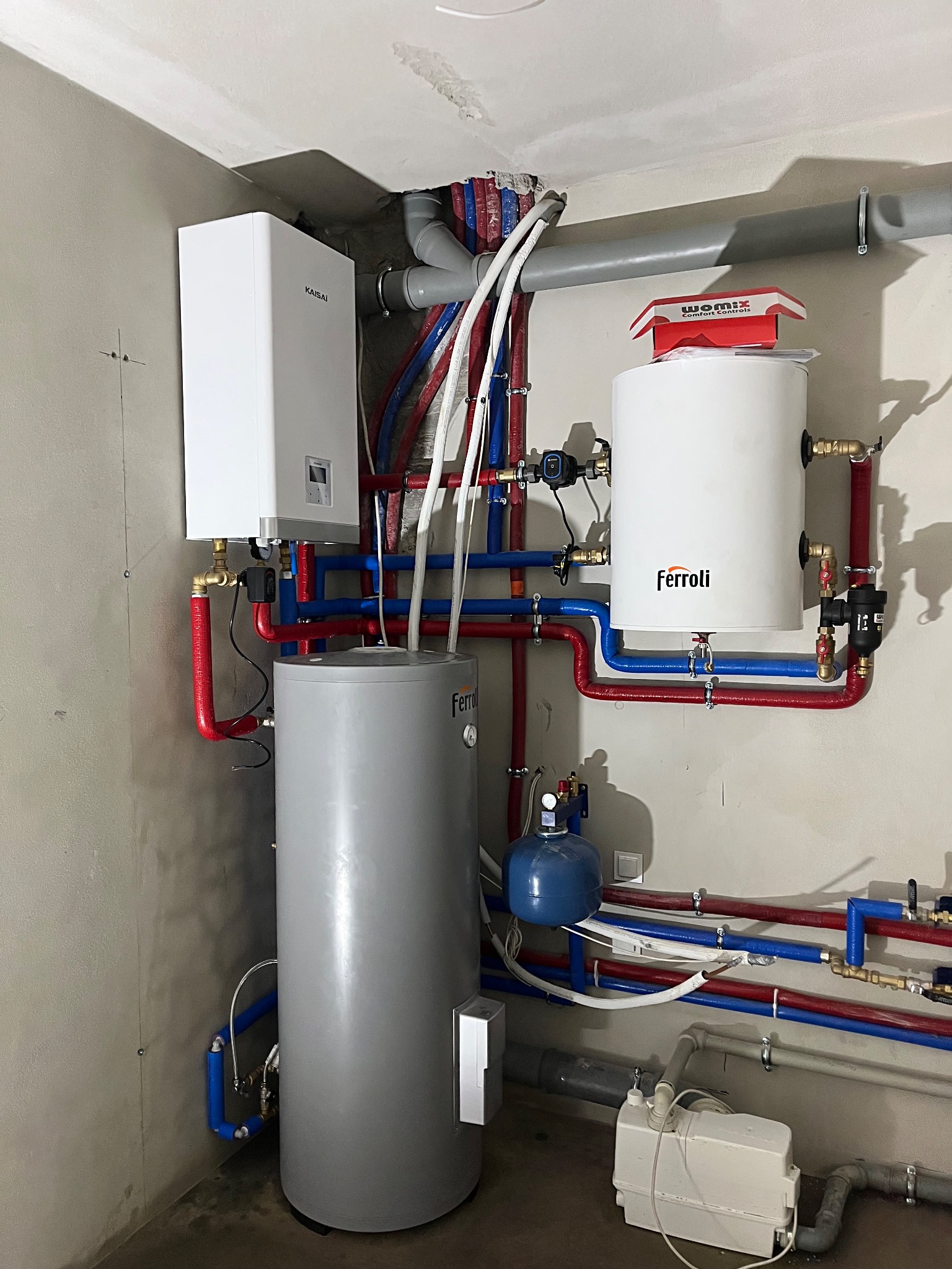 Zestaw urządzeń grzewczych: piec Kaisai, zbiorniki Ferroli i Womix, połączone rurami w kolorach czerwonym i niebieskim. Widoczne elementy instalacji hydraulicznej.