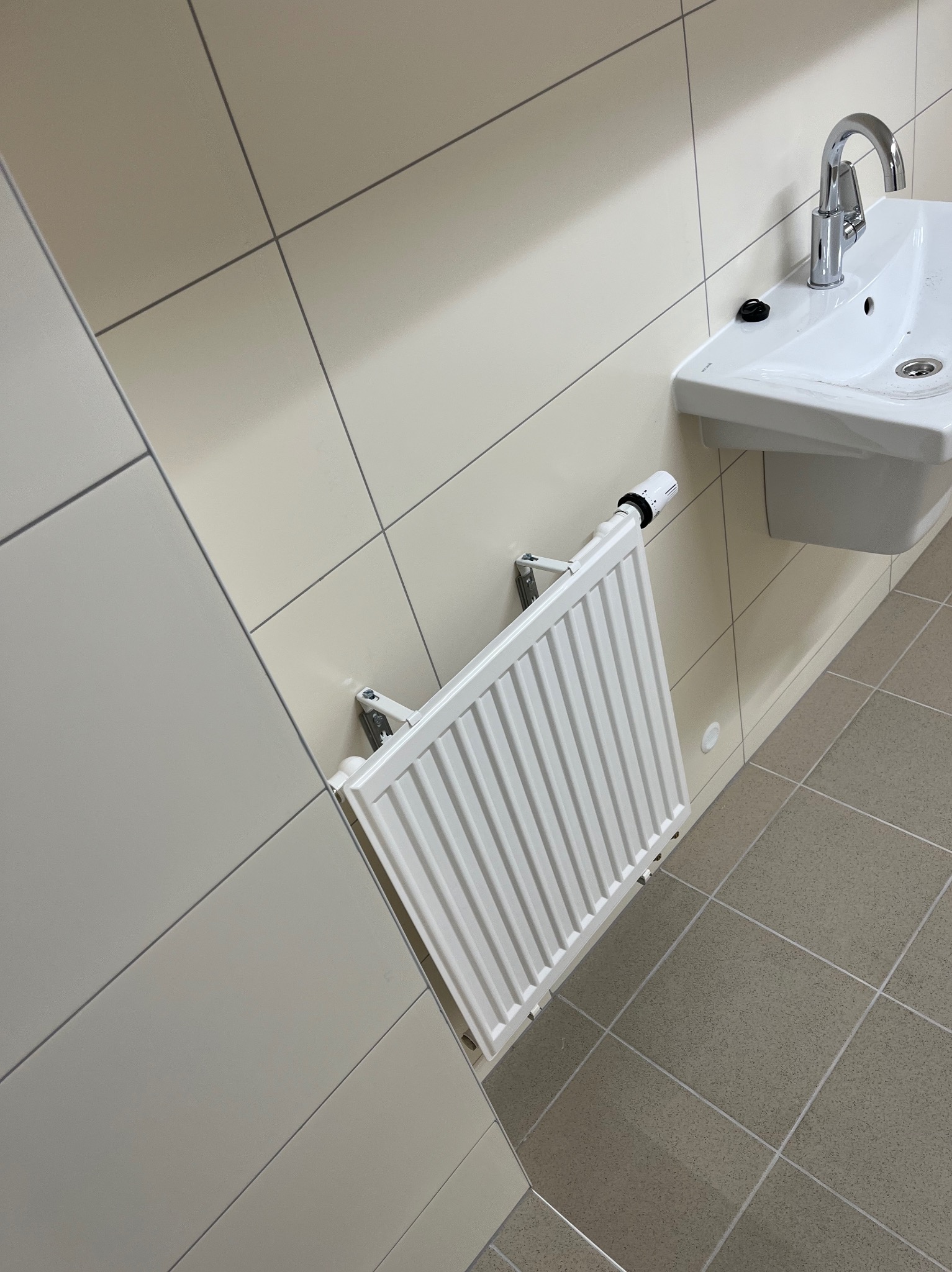 Biały grzejnik panelowy zamontowany na ścianie w łazience obok umywalki z chromowaną baterią, widok z dołu, płytki na ścianach i podłodze w odcieniach beżu.