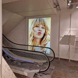 Firma budowlano-wykonczeniowa Gregor Grzegorz Małachowski - Prace remontowe w sklepie w Pruszczu Gdańskim: zabezpieczone schody ruchome z widocznym plakatem na ścianie i elementami wyposażenia sklepowego w tle.