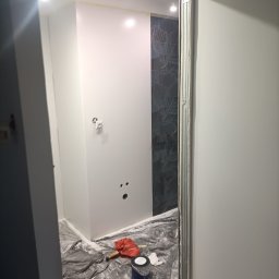 Firma budowlano-wykonczeniowa Gregor Grzegorz Małachowski - Świeżo pomalowane na biało ściany łazienki z widocznymi otworami pod instalacje, zabezpieczone podłogi, puszka z farbą i fragment ściany wykończonej płytkami z motywem roślinnym.