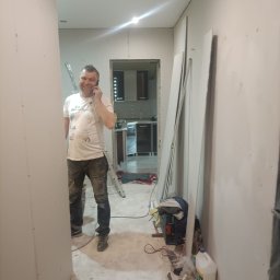 Firma budowlano-wykonczeniowa Gregor Grzegorz Małachowski - Uśmiechnięty mężczyzna rozmawia przez telefon w trakcie montażu płyt kartonowo-gipsowych w korytarzu, widoczne płyty, drabina i narzędzia.