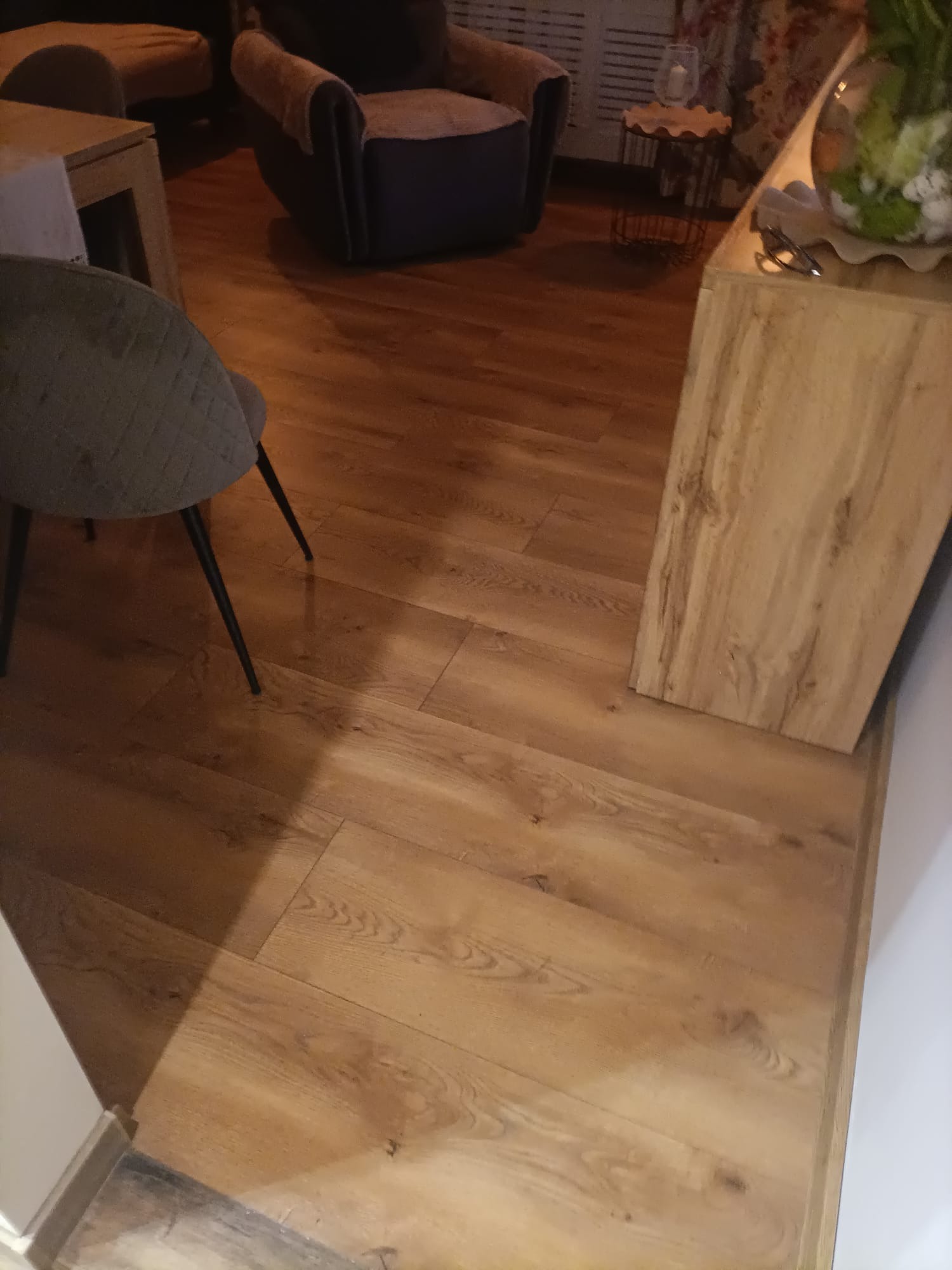 Ułożone panele podłogowe w odcieniach brązu, widoczne krzesło z szarą tapicerką i element szafki z jasnego drewna.