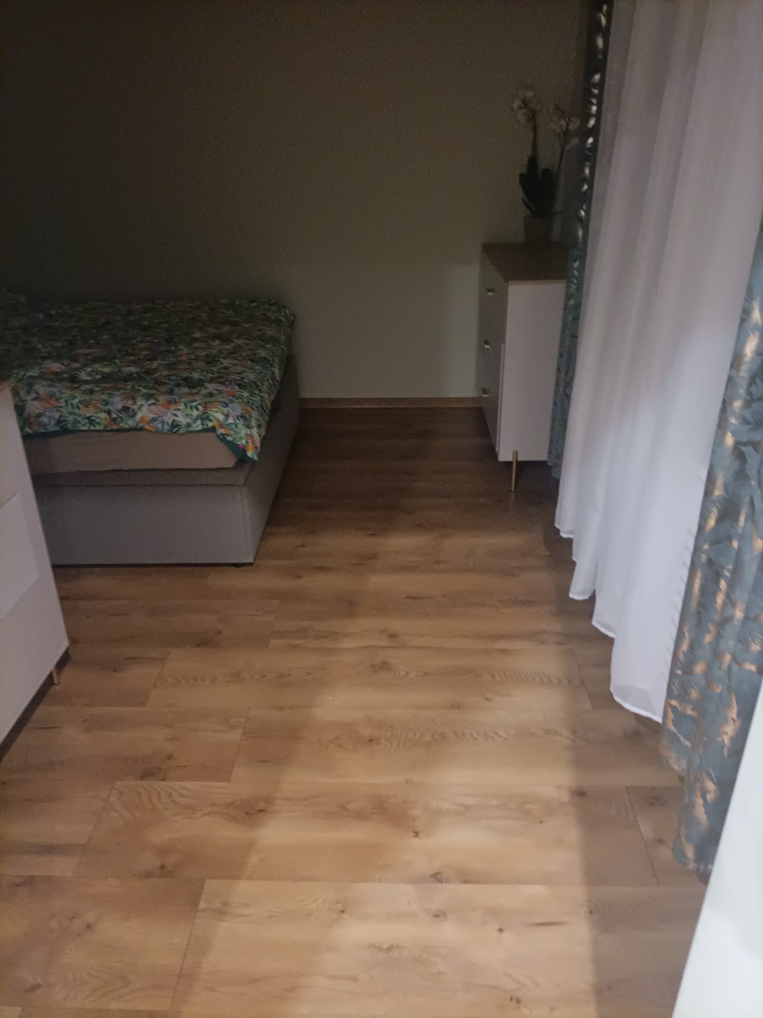 Jasne panele podłogowe w sypialni, widoczne fragment łóżka z wzorzystą pościelą, biała komoda z kwiatami i zwiewne, białe zasłony z delikatnym wzorem.