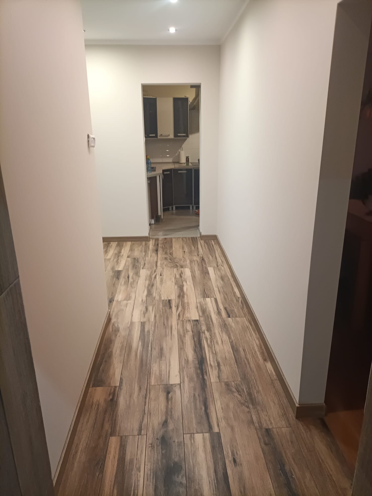 Korytarz z ułożonymi panelami podłogowymi imitującymi drewno, widok na kuchnię w tle.