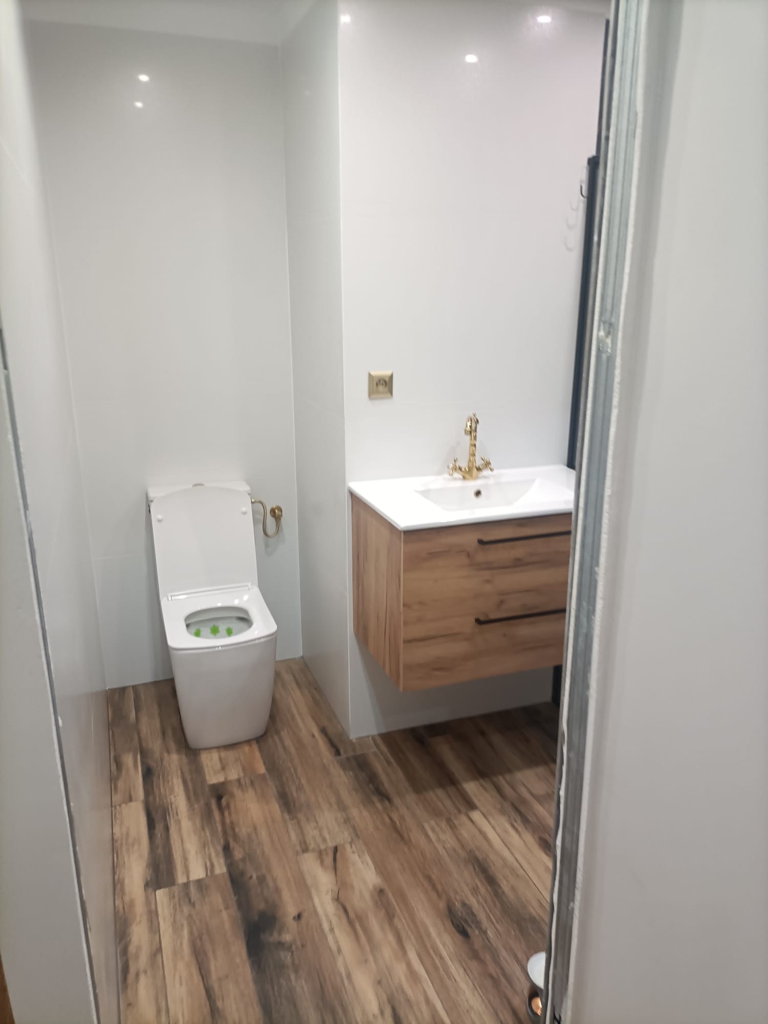 Nowoczesna łazienka z białymi płytkami ściennymi, drewnopodobną podłogą, wiszącą szafką z umywalką ze złotą armaturą i toaletą z zielonym odświeżaczem.