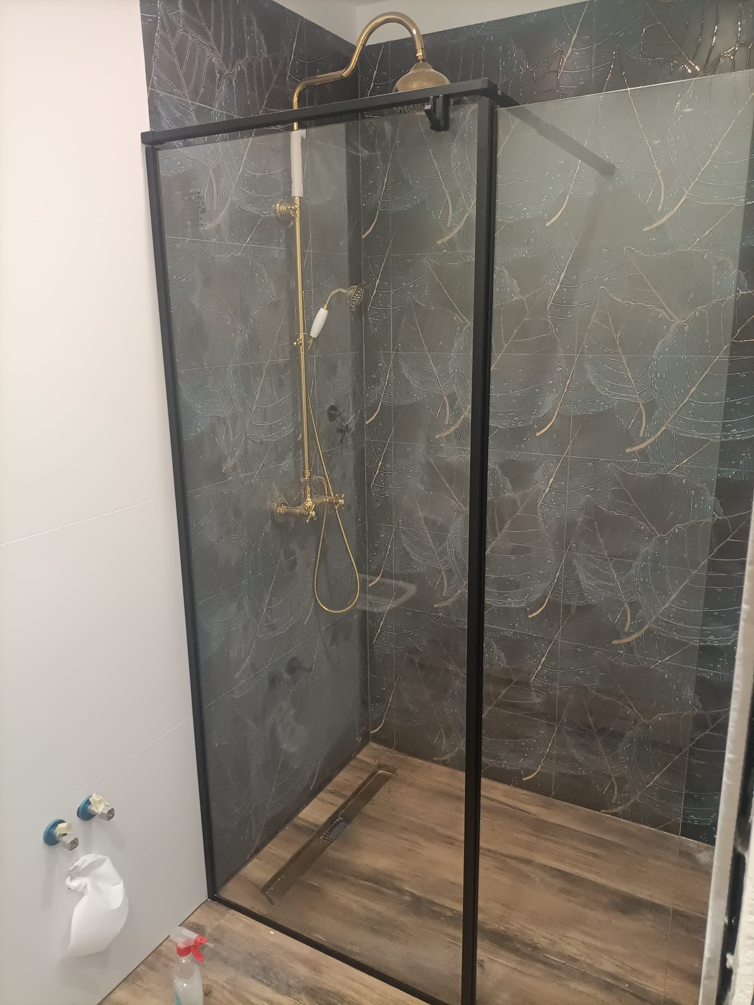 Kabina prysznicowa z czarną ramą i złotą armaturą na tle płytek z motywem liści, drewnopodobna podłoga z odpływem liniowym.