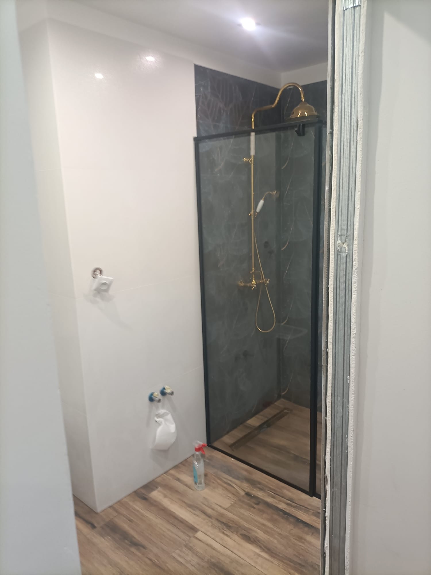 Częściowo wykończona łazienka z kabiną prysznicową w stylu retro, drewnopodobne panele podłogowe, białe ściany i widoczne elementy instalacji wodno-kanalizacyjnej.