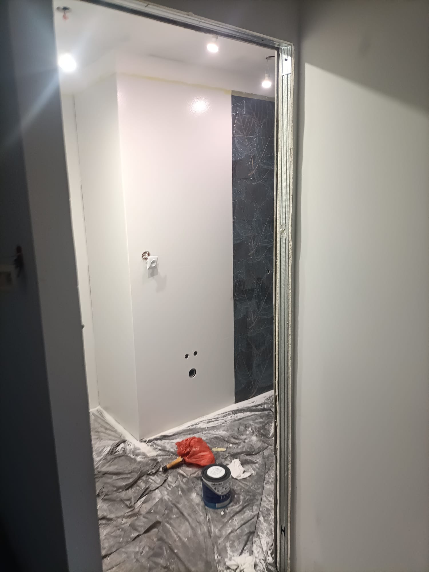 Świeżo pomalowane na biało ściany łazienki z widocznymi otworami pod instalacje, zabezpieczone podłogi, puszka z farbą i fragment ściany wykończonej płytkami z motywem roślinnym.