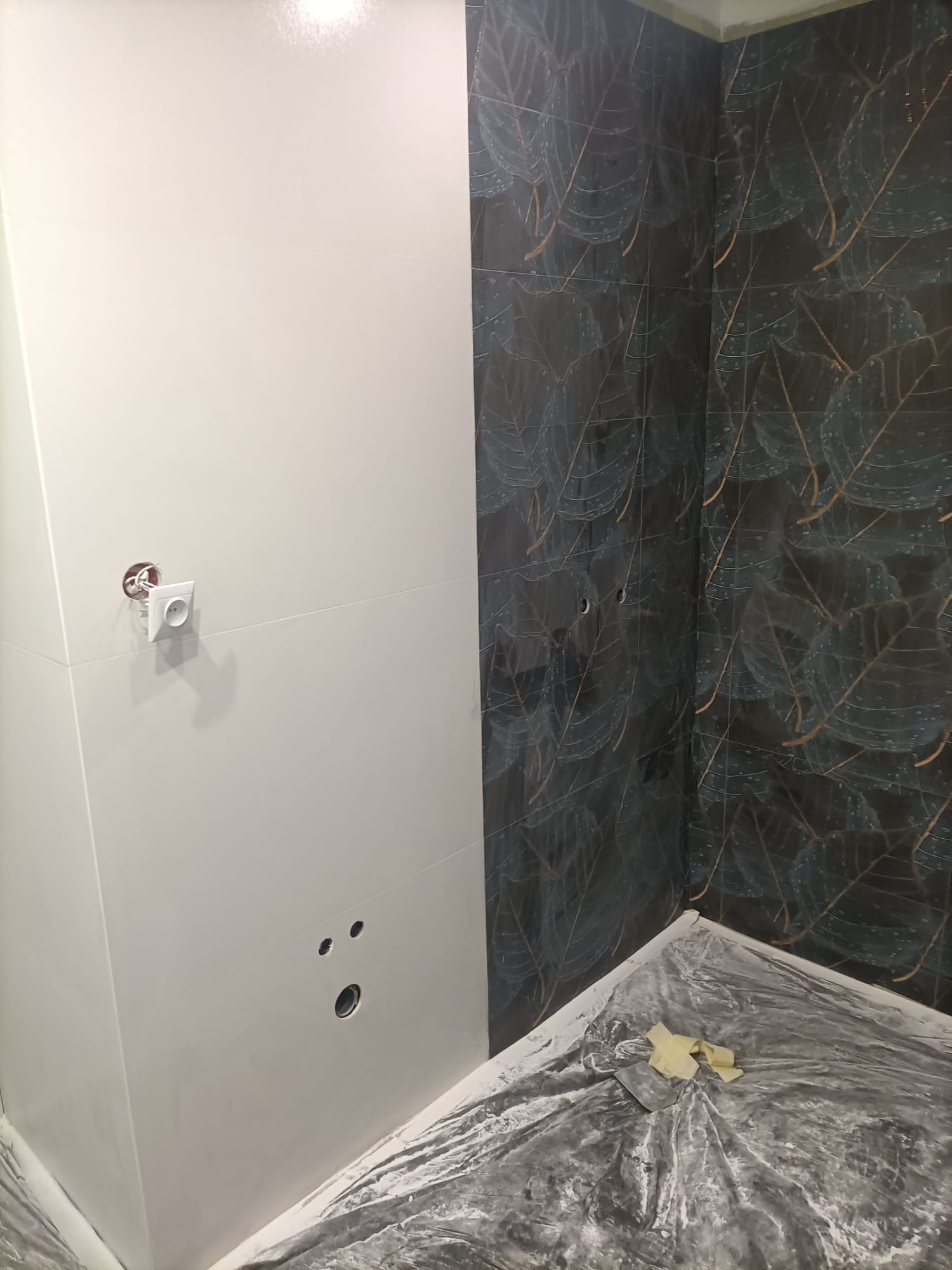 Świeżo wykafelkowana łazienka z białymi i wzorzystymi, ciemnymi płytkami, widoczne otwory na rury i gniazdka, podłoga zabezpieczona folią malarską.