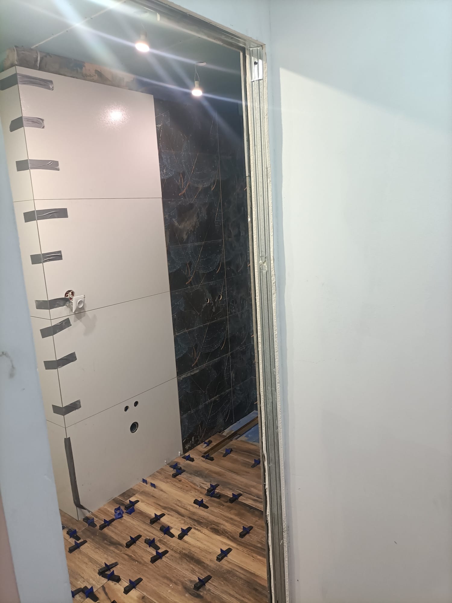 Łazienka w trakcie remontu: układanie płytek imitujących drewno na podłodze z użyciem klipsów poziomujących, częściowo wykończona ściana z jasnych płytek z szarą taśmą maskującą i fragment ściany...