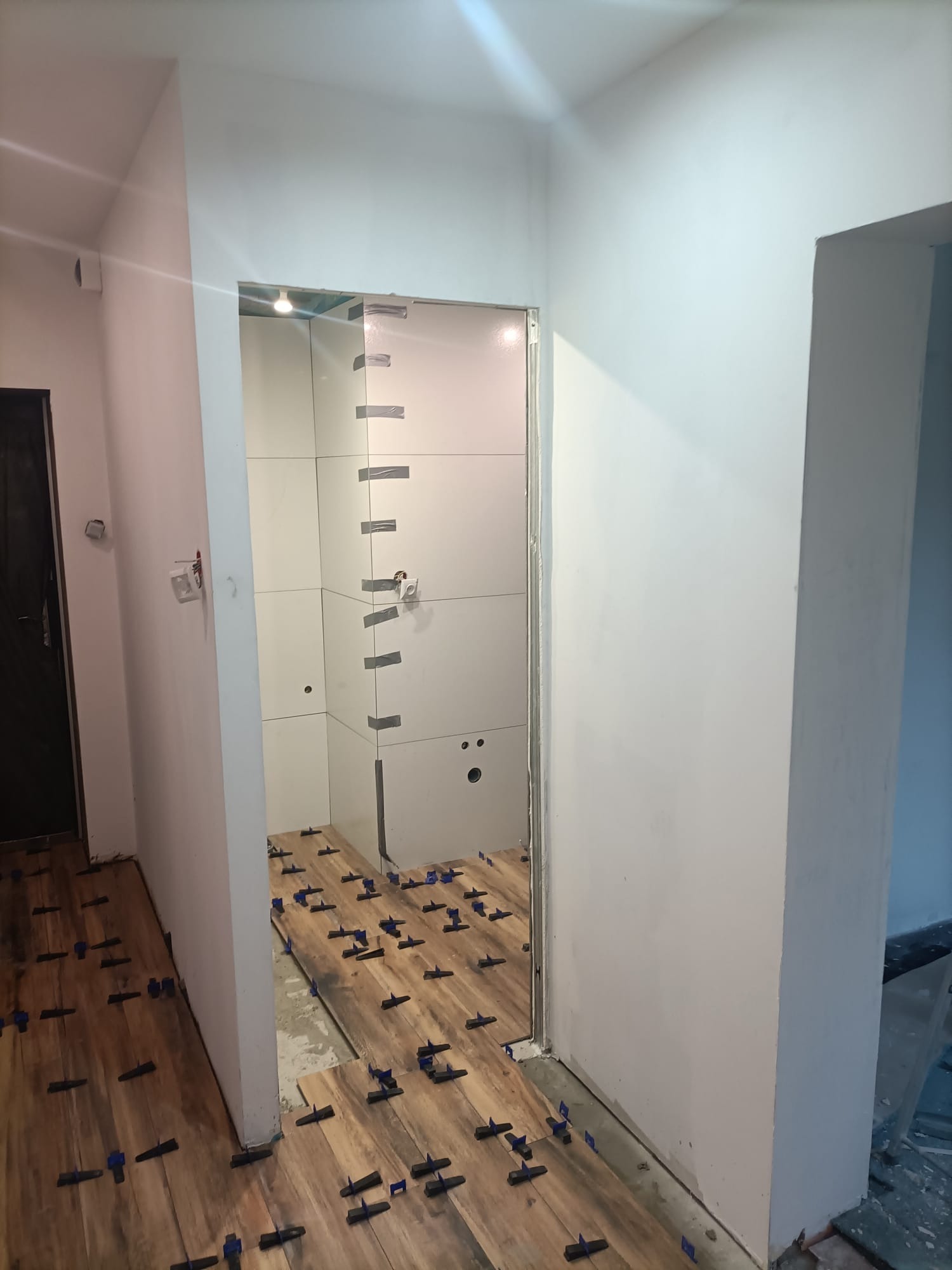 Montaż płytek imitujących drewno na podłodze w łazience, z użyciem niebieskich krzyżyków dystansowych, widoczne również płytki na ścianach zabezpieczone taśmą malarską.