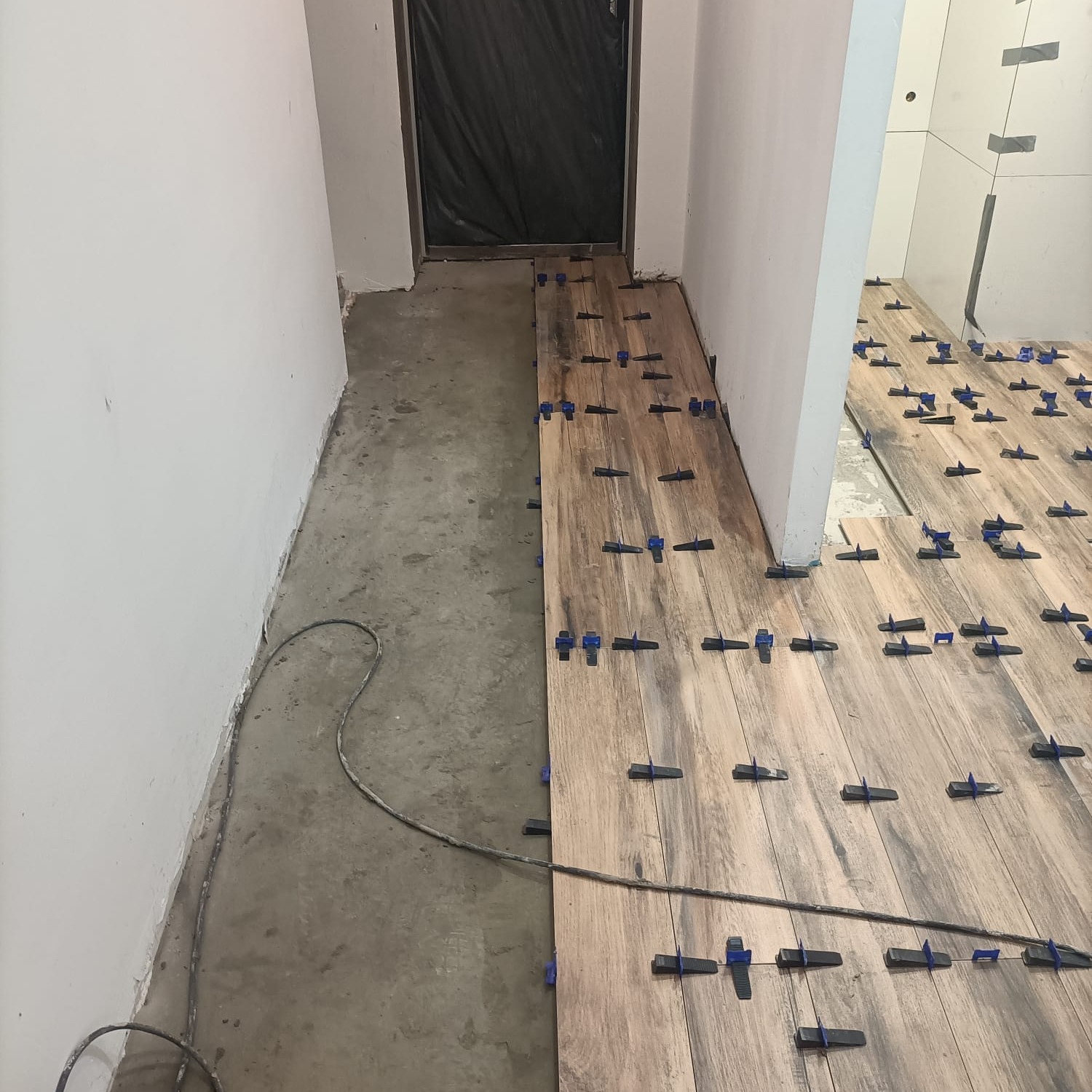 Proces układania podłogi z płytek imitujących drewno, z użyciem niebieskich klinów poziomujących, widoczne surowe ściany i podłoże w trakcie remontu.