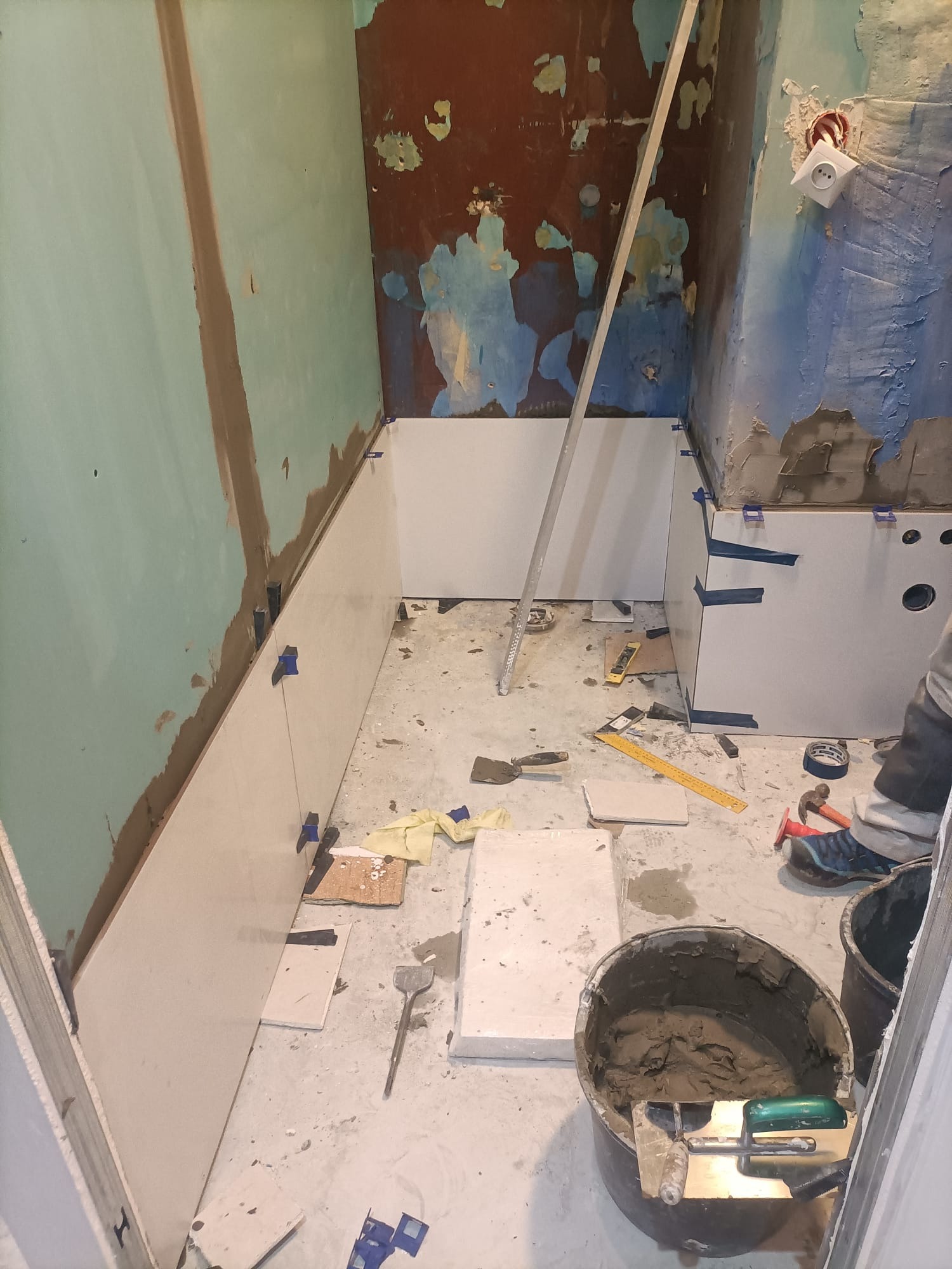 Przygotowanie do układania białych płytek w małej łazience z nierównymi ścianami; widoczne narzędzia, wiadro z zaprawą i fragment nogi glazurnika