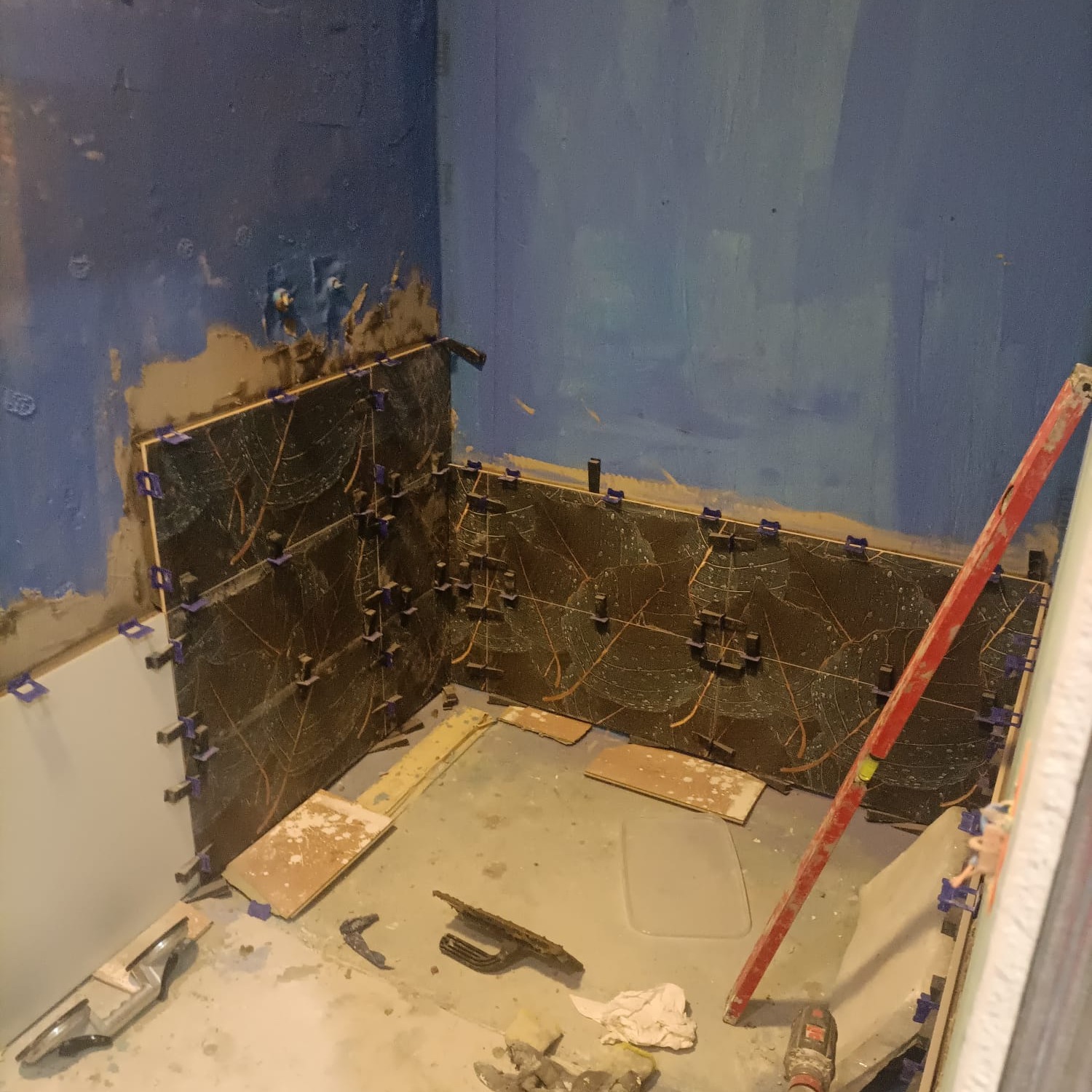 Fragment łazienki w trakcie prac: ułożone ciemne płytki z wzorem, poziomica oparta o ścianę, niebieskie ściany w stanie surowym, widoczne narzędzia glazurnicze.