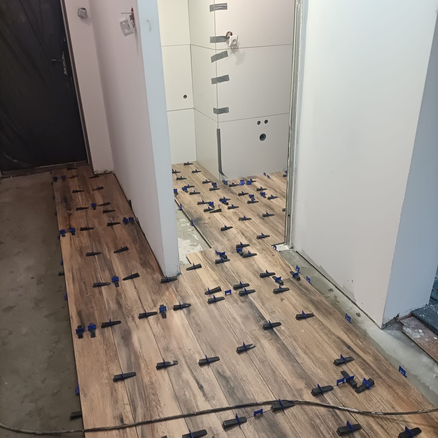 Układanie drewnopodobnych płytek podłogowych z użyciem systemów poziomowania, widoczne fragmenty kabla i przygotowywanej ściany w łazience.