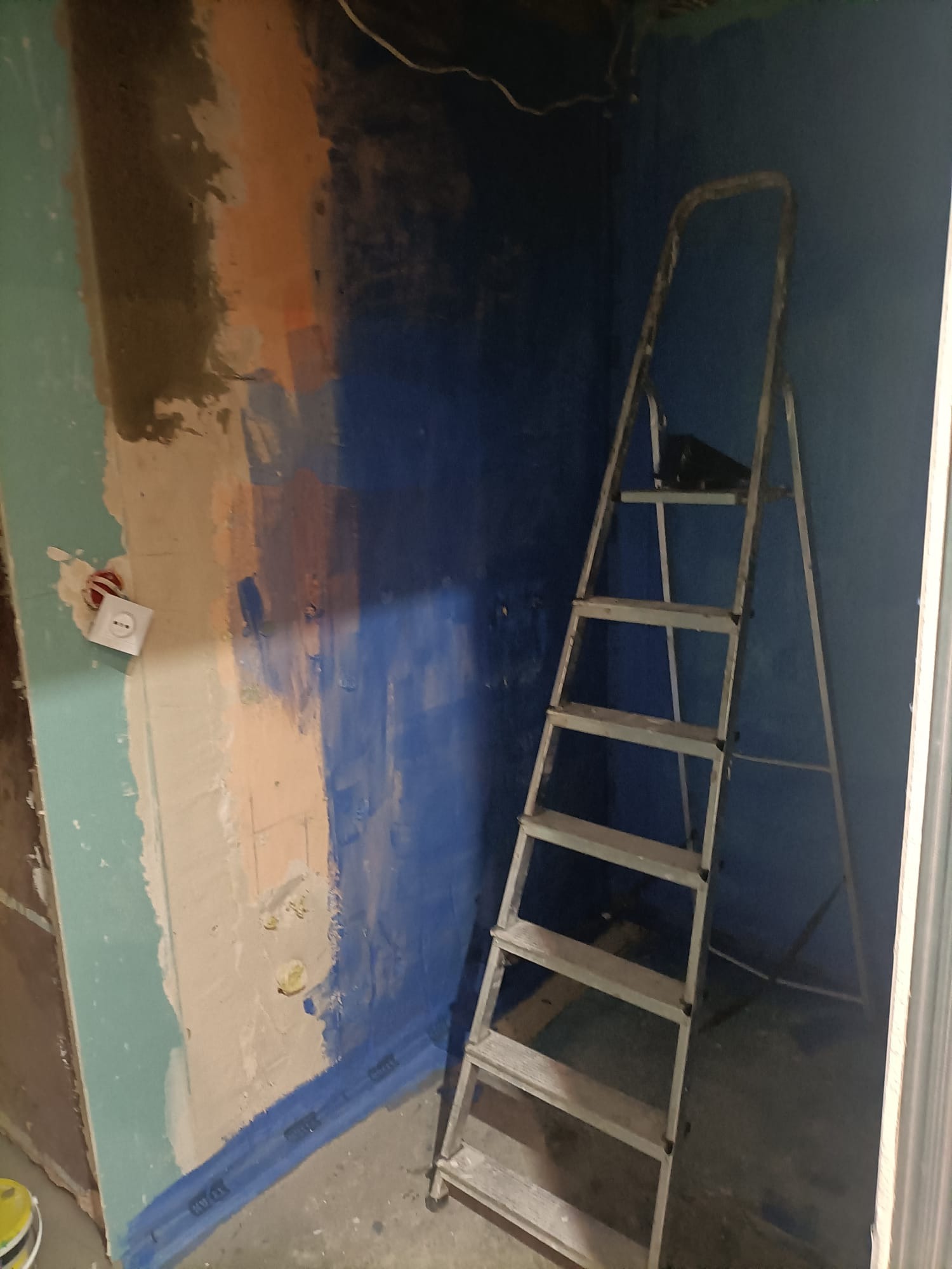 Ściana w trakcie przygotowania do prac wykończeniowych, widoczne fragmenty gładzi, niebieska farba gruntująca i drabina przystawna.
