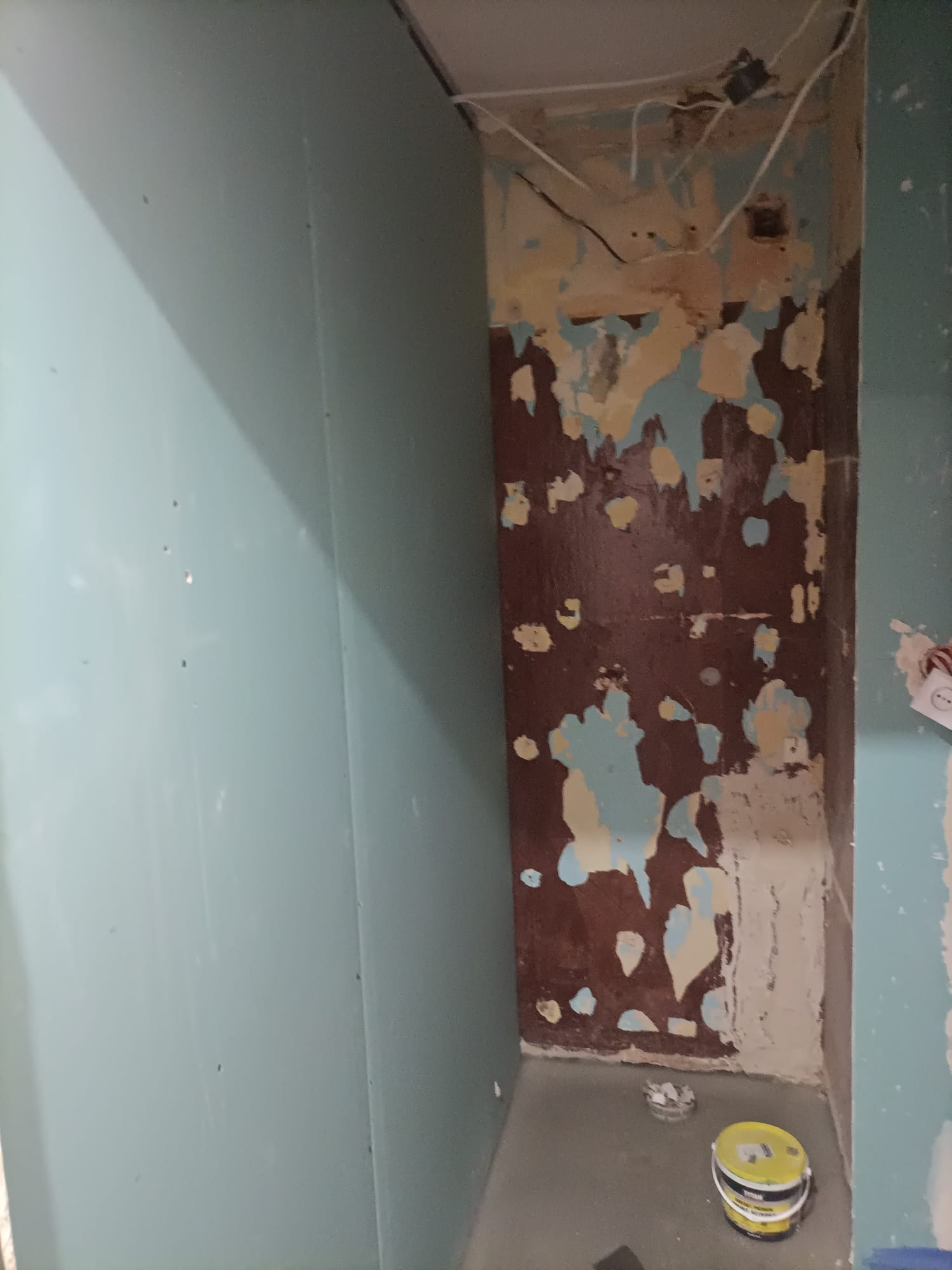 Przygotowanie ściany do prac wykończeniowych: płyta gipsowo-kartonowa i fragment ściany ze starą farbą oraz resztkami gładzi, widoczne przewody elektryczne, na podłodze puszka z masą szpachlową.