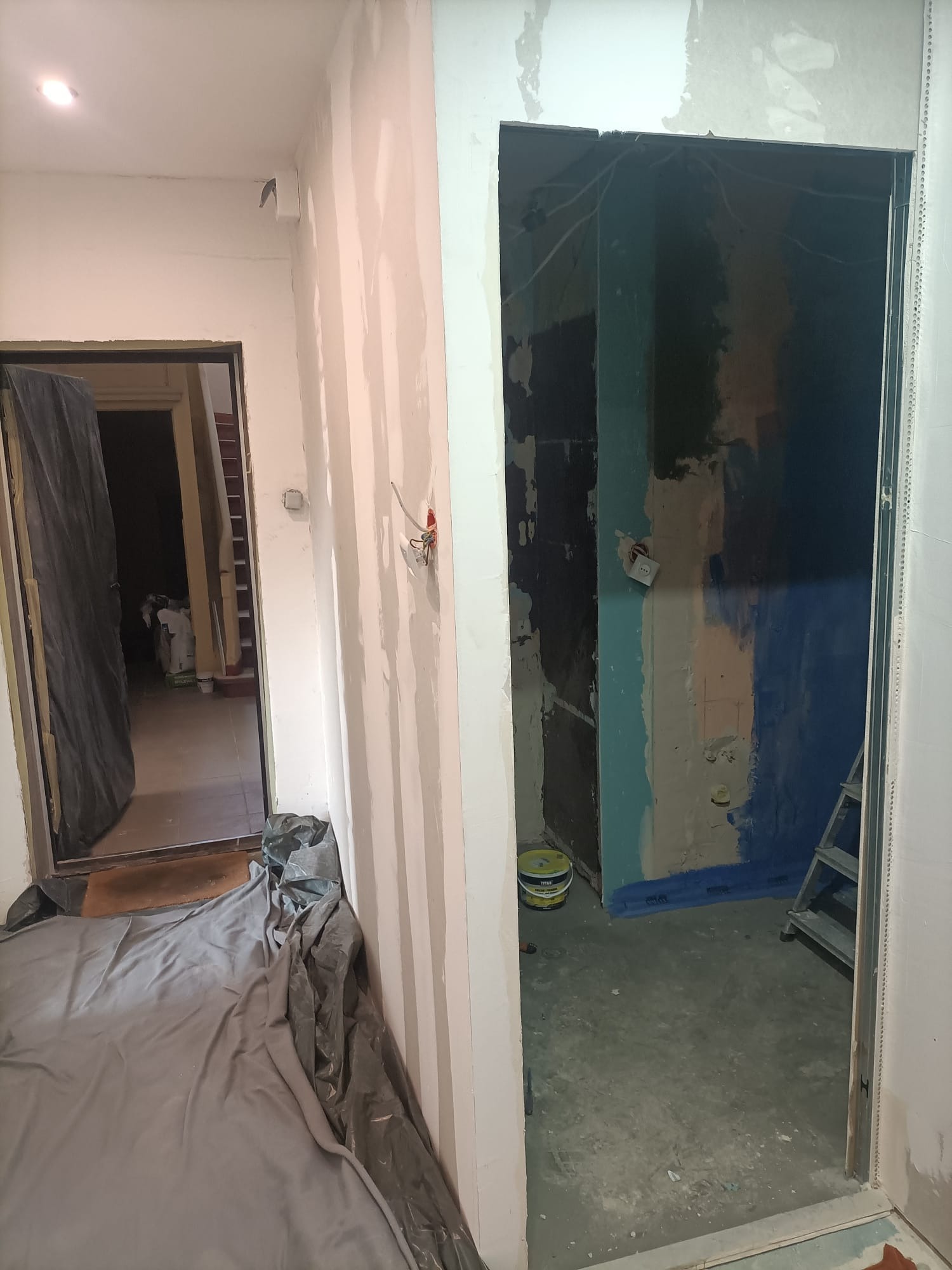 Wnętrze w trakcie remontu: przygotowanie ścian pod gładzie, zabezpieczona podłoga folią malarską, widoczne kable elektryczne, drabina i wiadro z farbą w tle.