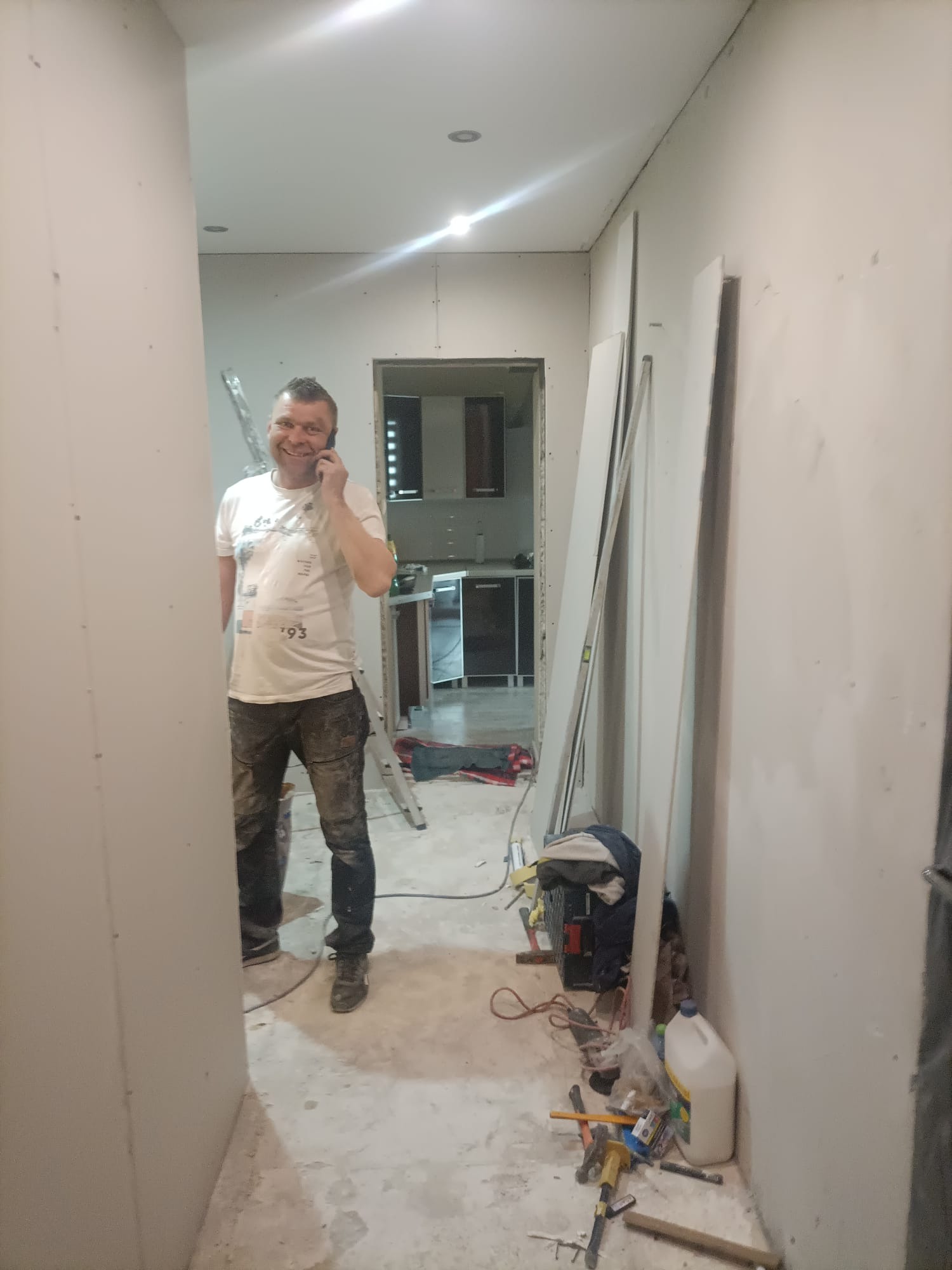 Uśmiechnięty mężczyzna rozmawia przez telefon w trakcie montażu płyt kartonowo-gipsowych w korytarzu, widoczne płyty, drabina i narzędzia.