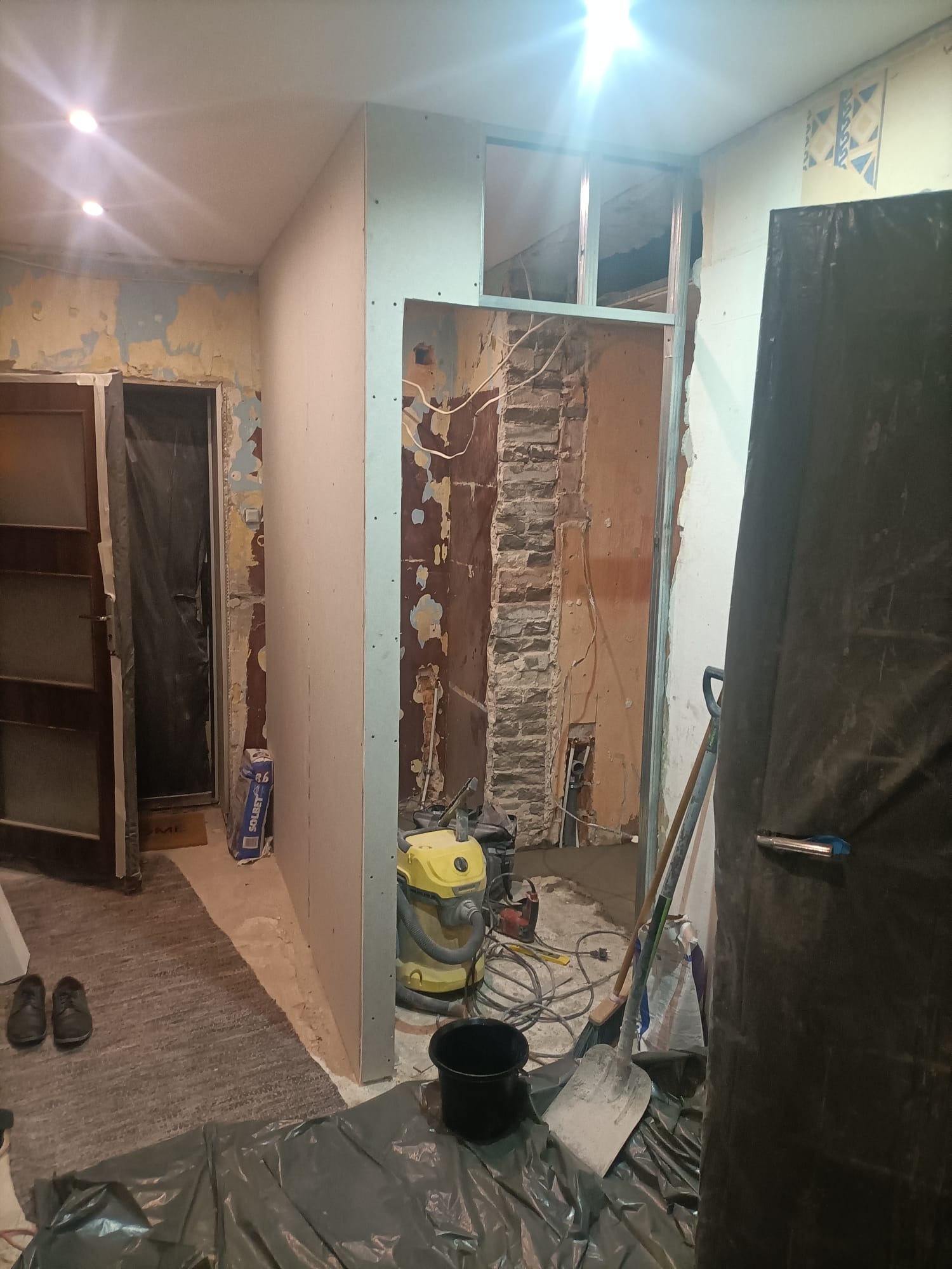 Wnętrze w trakcie remontu: widoczne odsłonięte cegły, fragment nowej ścianki działowej z płyt kartonowo-gipsowych, żółty odkurzacz przemysłowy, narzędzia, zabezpieczona folią podłoga i fragmenty...