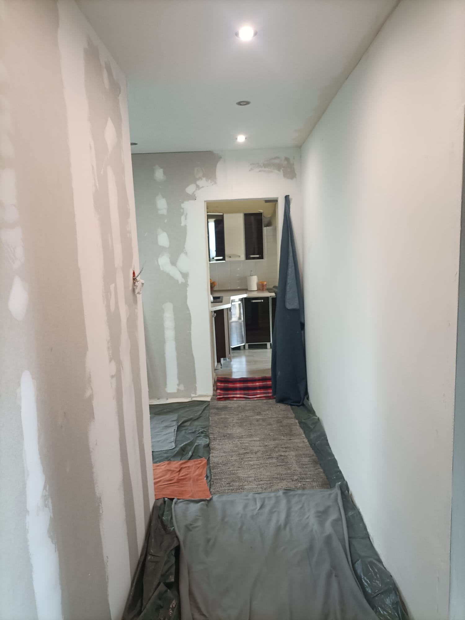 Korytarz w trakcie prac wykończeniowych; ściany częściowo pokryte gładzią, podłoga zabezpieczona folią malarską, widoczny fragment kuchni w tle.