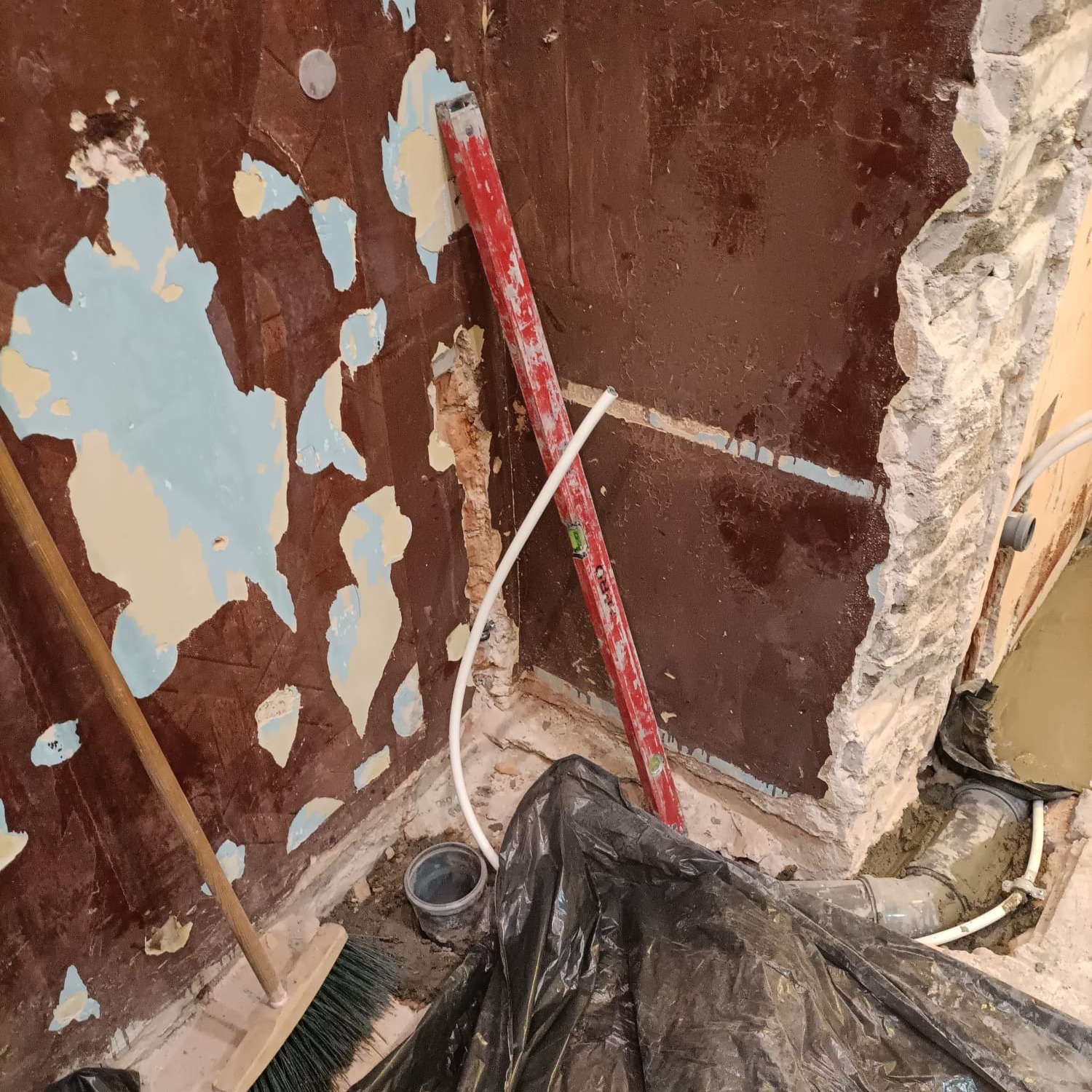 Fragment ściany w trakcie remontu łazienki: zerwana glazura, odsłonięta cegła, widoczny poziomica, rury instalacyjne, zanieczyszczenia i resztki materiałów budowlanych.