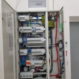 Otwarta szafa elektryczna z precyzyjnie ułożonymi kablami o różnych kolorach, widoczne bezpieczniki i schemat na drzwiach, schludne wykonanie.