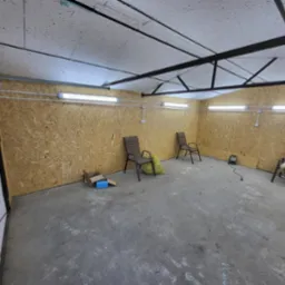Wnętrze garażu z lampami jarzeniowymi na ścianach z płyty OSB. Na podłodze widoczne krzesła i drobne przedmioty. Sufit z widoczną konstrukcją stalową.