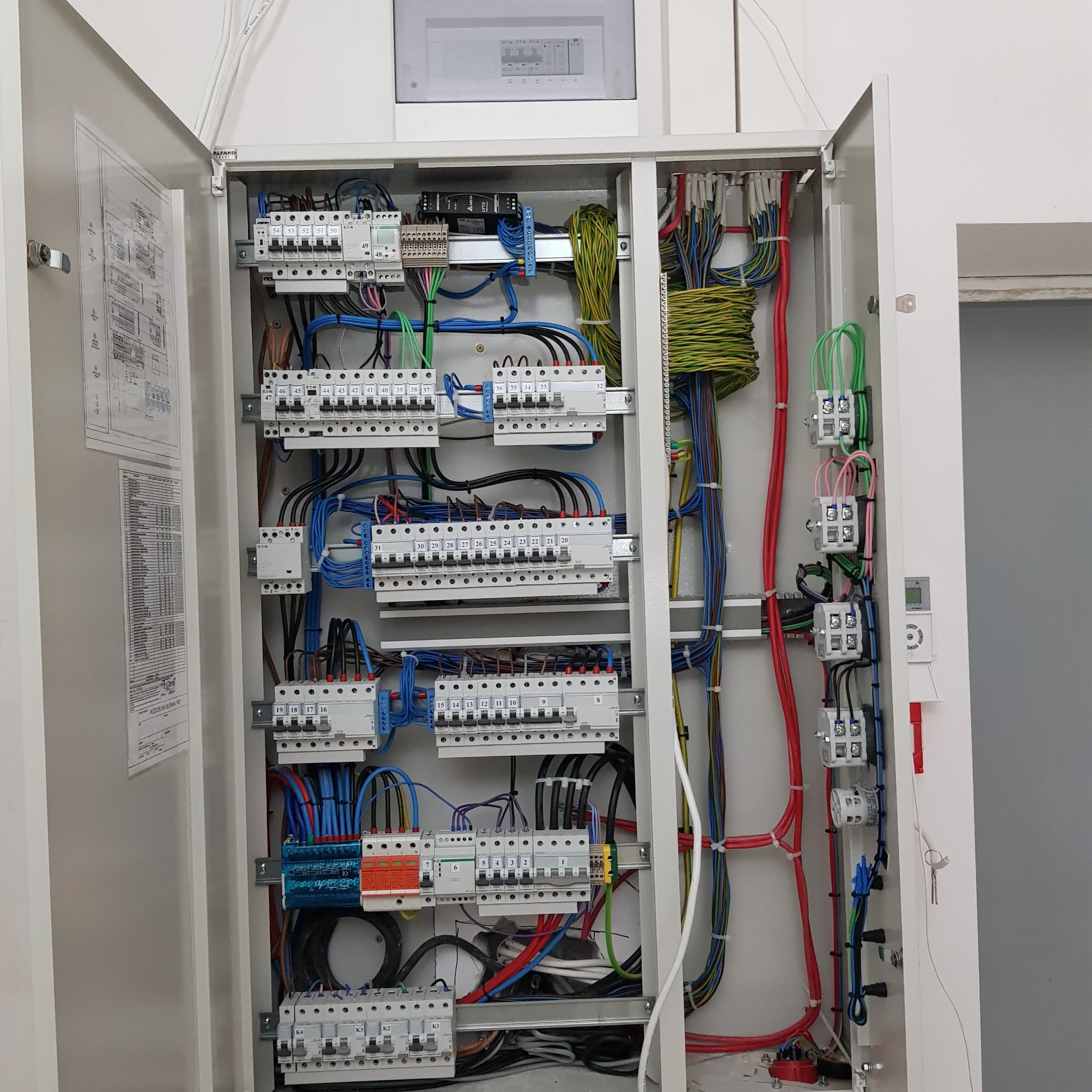 Otwarta szafa elektryczna z precyzyjnie ułożonymi kablami o różnych kolorach, widoczne bezpieczniki i schemat na drzwiach, schludne wykonanie.