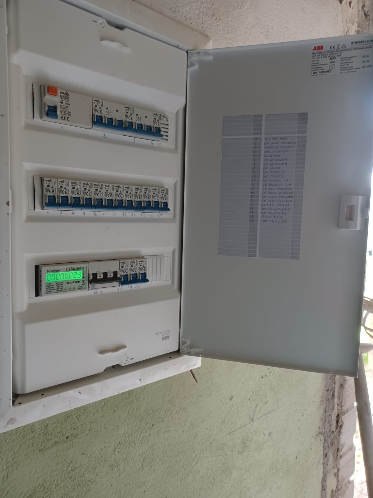 Otwarta skrzynka elektryczna ABB z bezpiecznikami i licznikiem Schelinger na ścianie. Widoczne oznaczenia obwodów na wewnętrznej stronie drzwiczek.