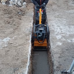 MIA BARTOSZ SOKOŁOWSKI - Pracownik w odblaskowej kamizelce zagęszcza wąski wykop ubijarką wibracyjną, w tle widoczna minikoparka SANY i gruz.