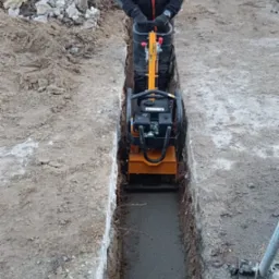 Pracownik w odblaskowej kamizelce zagęszcza wąski wykop ubijarką wibracyjną, w tle widoczna minikoparka SANY i gruz.