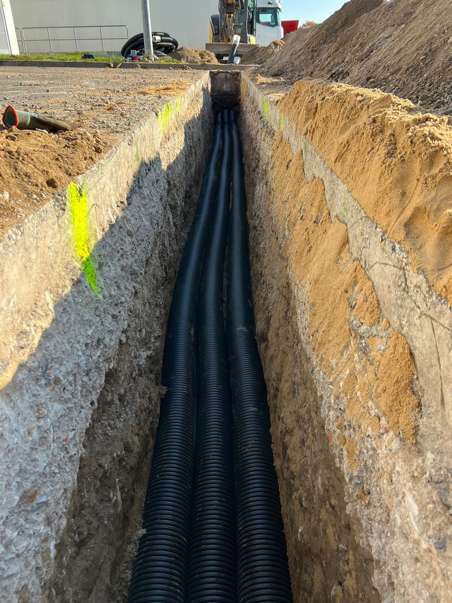 Wąski wykop z trzema czarnymi rurami karbowanymi w ziemi, widoczny fragment żółtej farby na ścianie wykopu, w tle koparka i budynek.