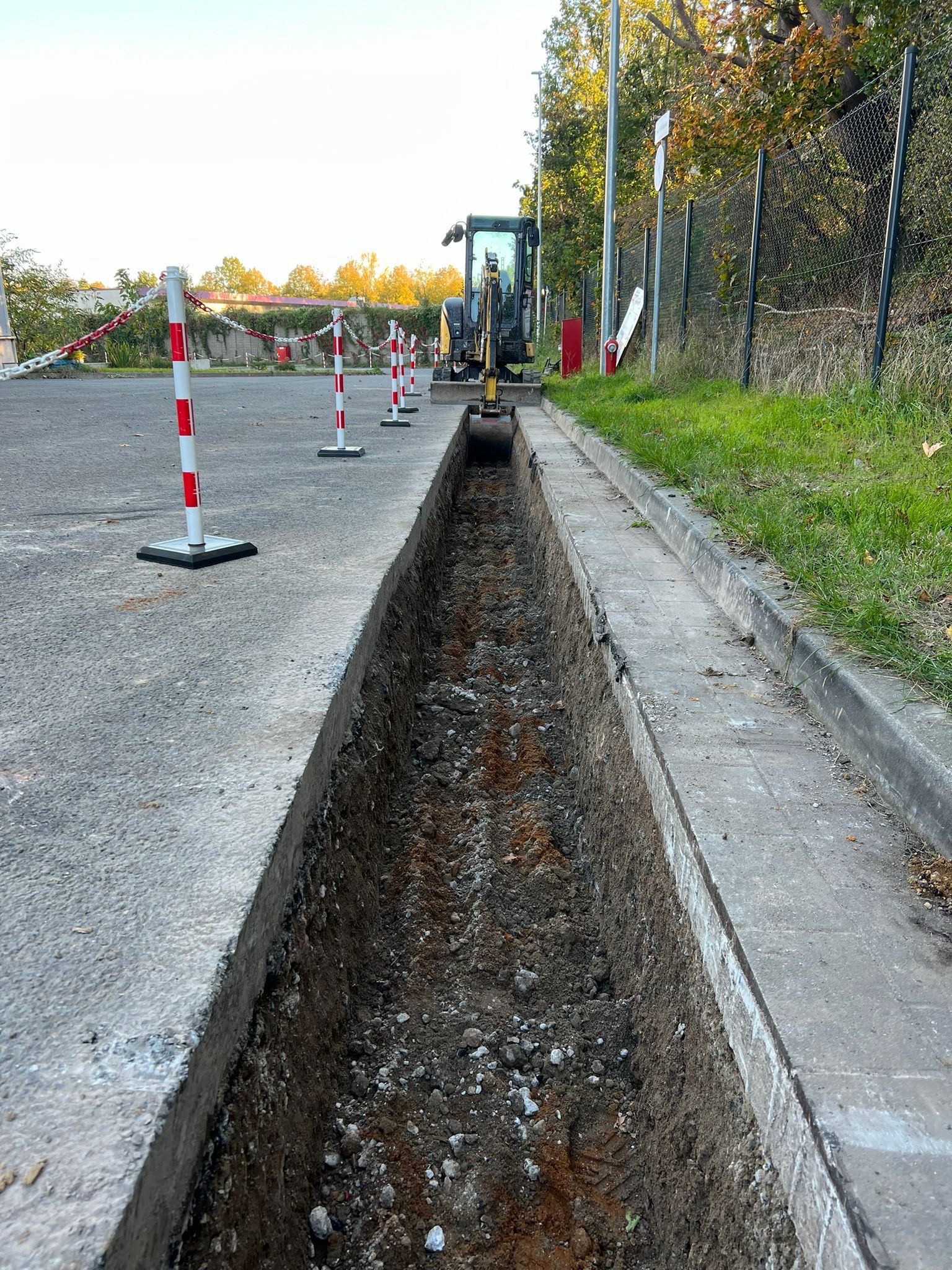 Długi wykop wzdłuż betonowej drogi, zabezpieczony słupkami z łańcuchem, z widoczną w oddali małą koparką, sugerujący prace instalacyjne.