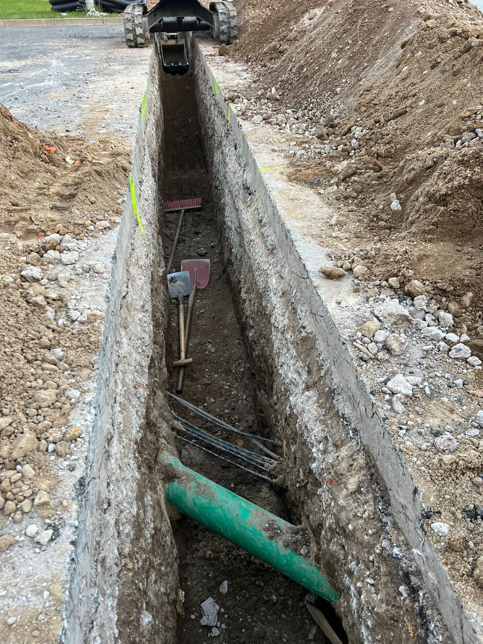 Wąski wykop w ziemi z widoczną zieloną rurą, kablami, łopatą i miotłą. Na górze kadru widoczna minikoparka.