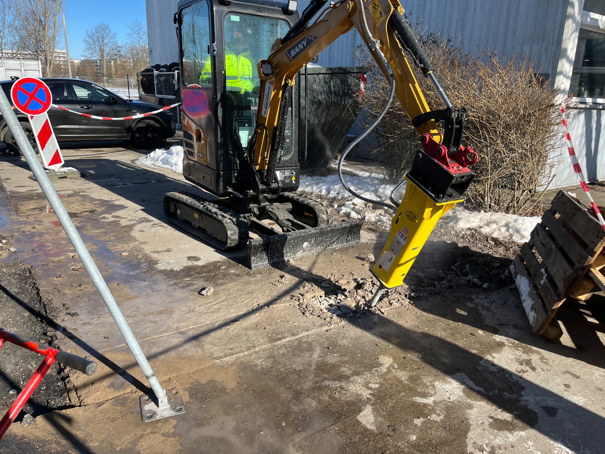 Żółta minikoparka z młotem pneumatycznym kruszy betonową nawierzchnię; widoczne ślady pracy, gruz i zaparkowany samochód w tle.