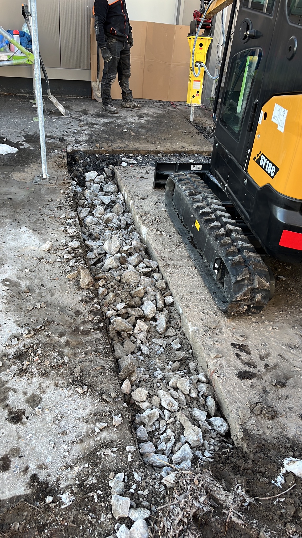 Wąski wykop w betonie wypełniony gruzem, obok gąsienica minikoparki i pracownik w roboczym ubraniu, w tle ściana budynku i młot pneumatyczny.