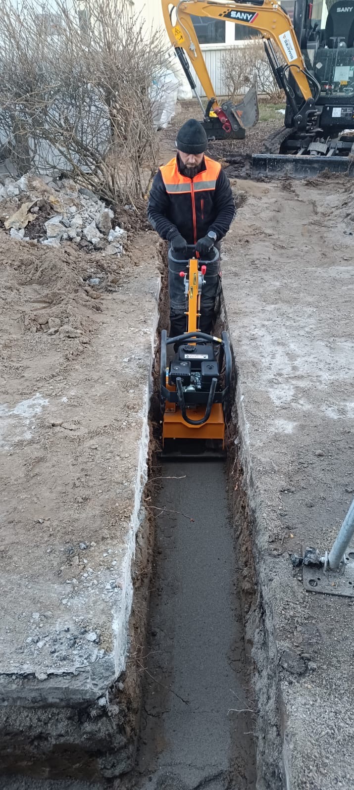 Pracownik w odblaskowej kamizelce zagęszcza wąski wykop ubijarką wibracyjną, w tle widoczna minikoparka SANY i gruz.