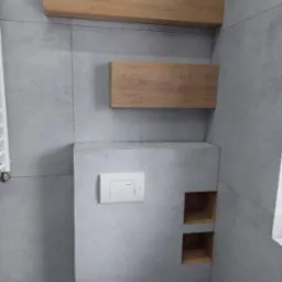 Szara łazienka z podtynkową toaletą, drewnianymi półkami nad nią i dwoma wnękami na ścianie wykończonej płytkami imitującymi beton.