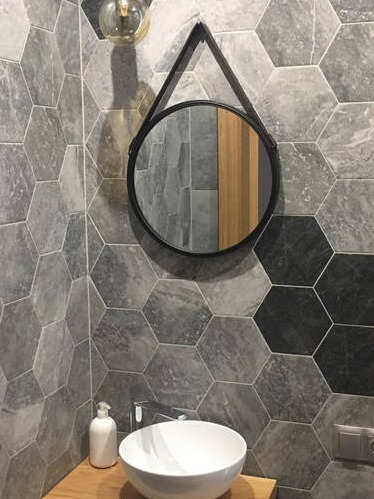 Łazienka z heksagonalnymi płytkami w odcieniach szarości i czerni, okrągłe lustro z czarną ramą na skórzanym pasku, biała umywalka nablatowa, szklana lampa sufitowa z widoczną żarówką.