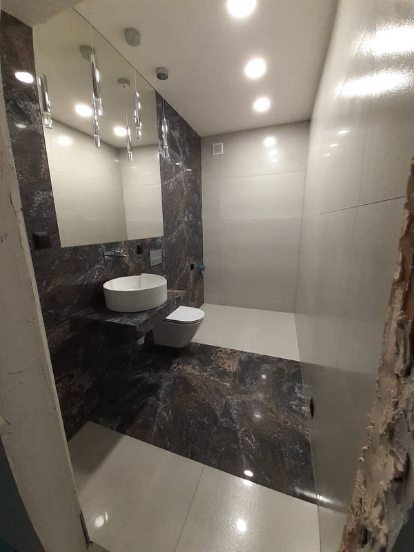 Nowoczesna łazienka z ciemnym marmurem na ścianie i podłodze, biała umywalka nablatowa i toaleta, lustro z wiszącymi lampami, oświetlenie sufitowe punktowe.